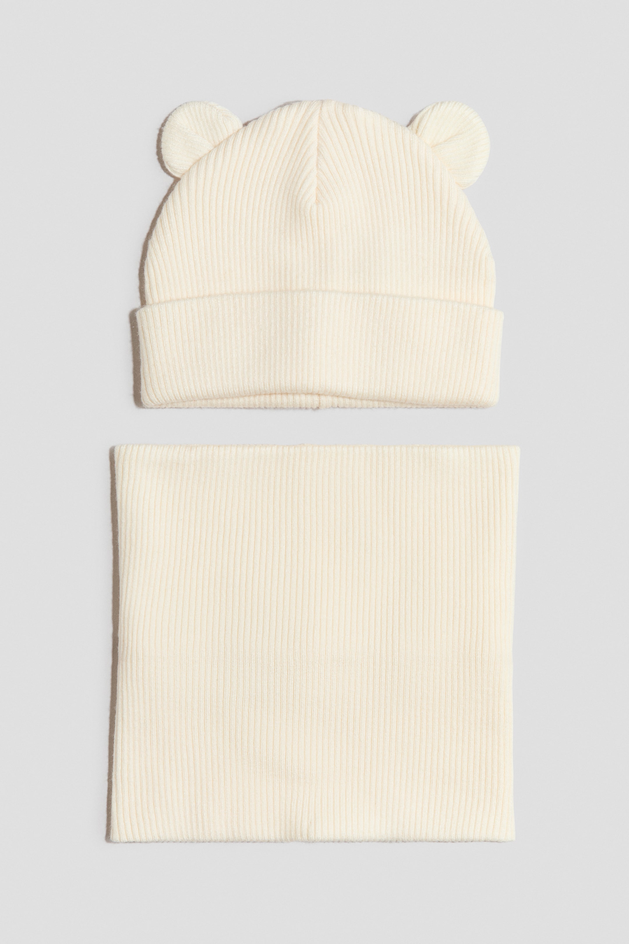 View larger image: A cream-coloured ribbed knit hat features two small, rounded ear-like protrusions on its crown and a wide, folded brim. A matching rectangular ribbed knit scarf lies flat below it.