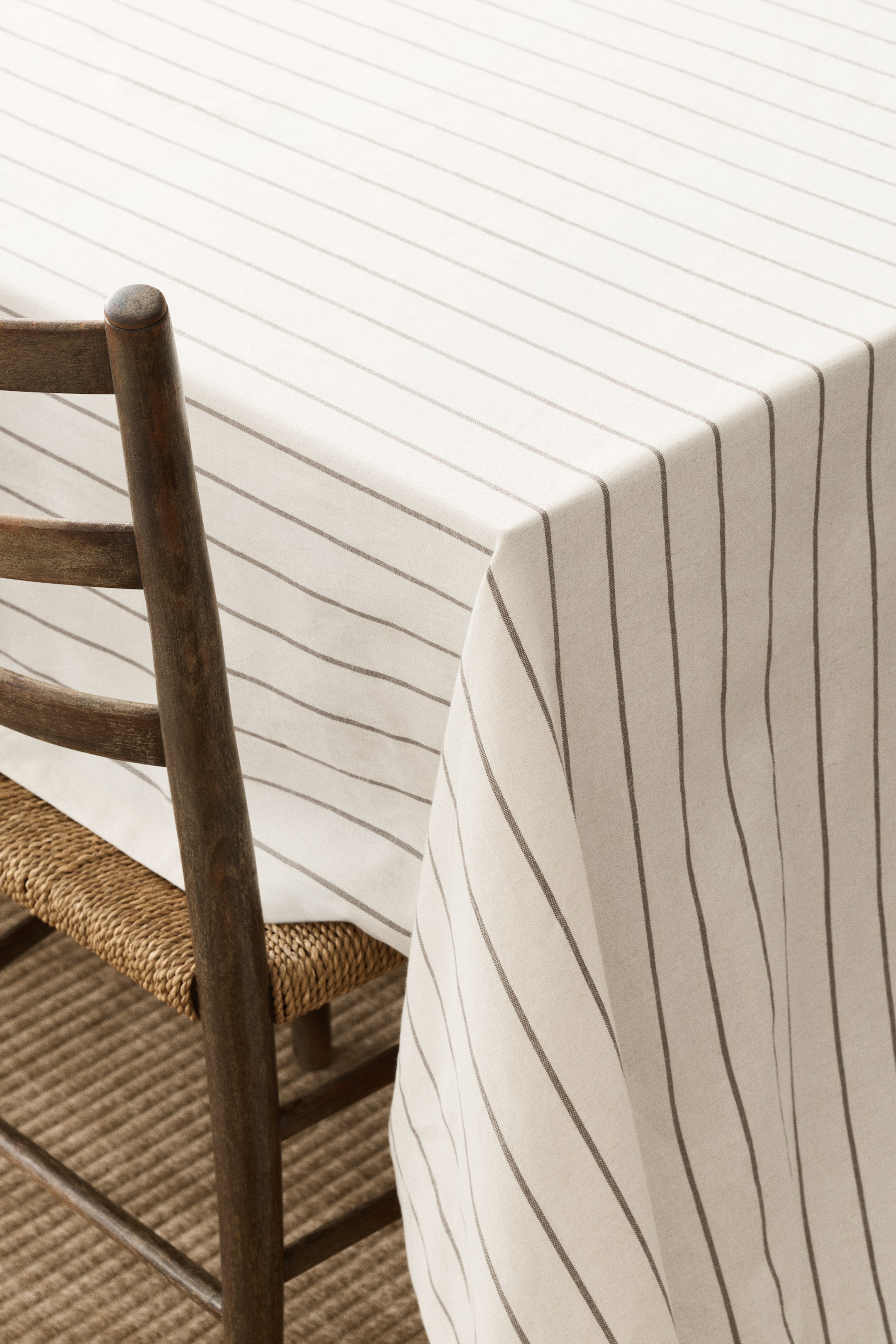 Voir une image plus grande: Une nappe blanc cassé avec de fines rayures gris foncé recouvre une table. Les rayures s'étendent horizontalement sur le dessus de la table et verticalement le long des côtés drapés, et une chaise en bois cache partiellement le tissu.