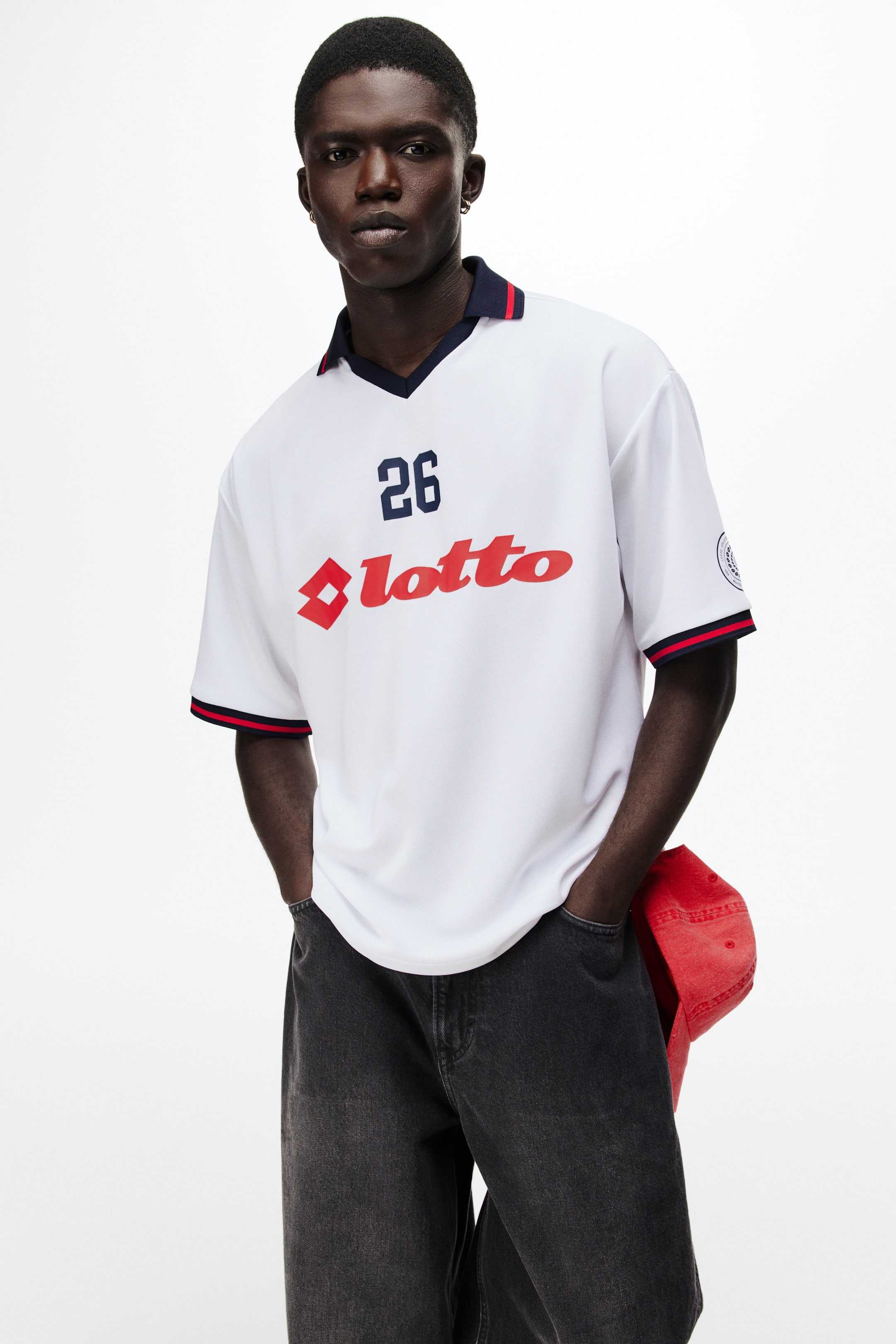 View larger image: A white short-sleeved jersey featuring a navy blue V-neck collar with red and white stripe detailing. The short sleeves are accented with navy blue, red, and white striped cuffs, and a circular patch is present on the left sleeve. The front displays the number "26" in navy blue alongside a red "lotto" logo with a diamond graphic.