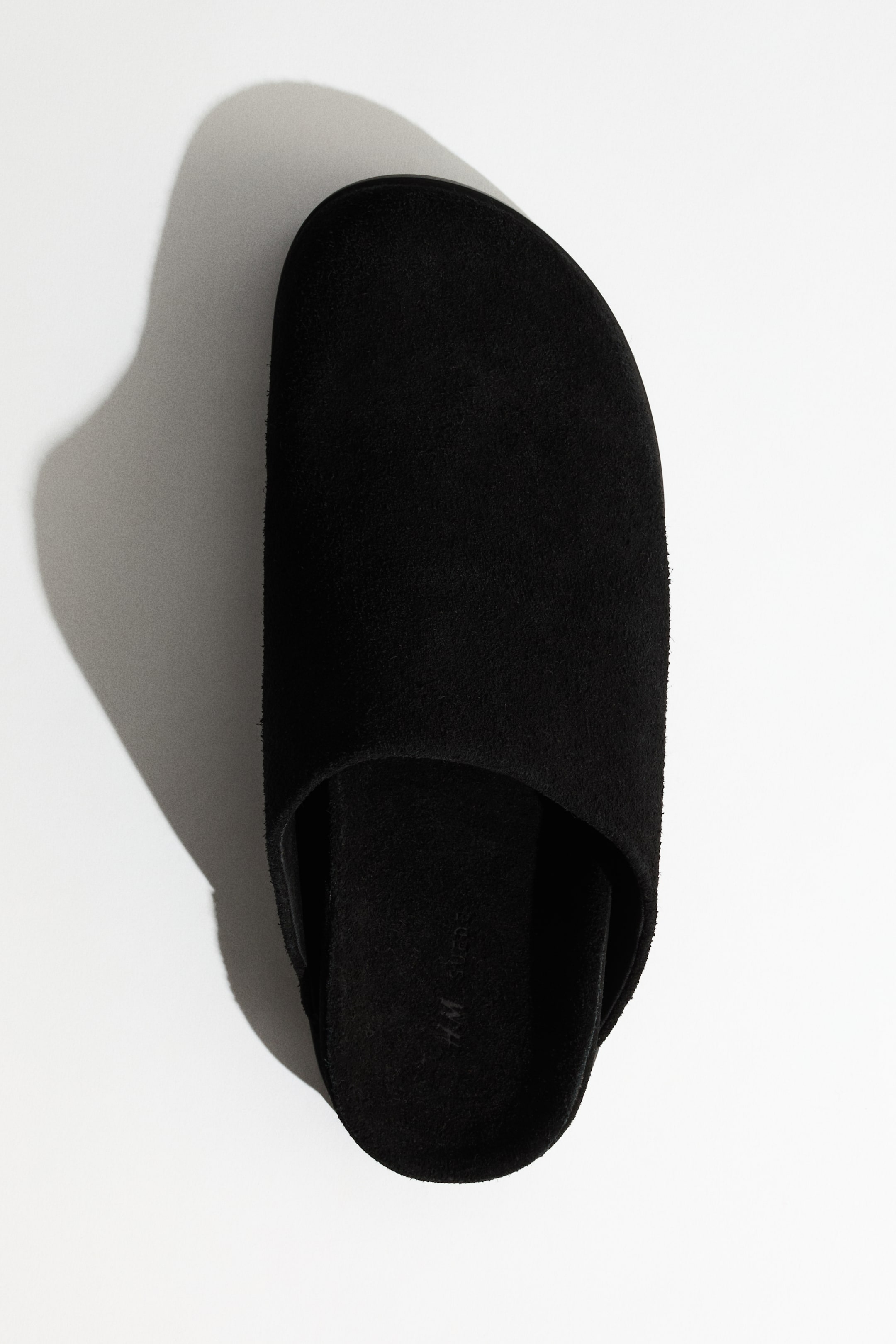 Ampliar la imagen: Una pantufla de gamuza negra está orientada ligeramente hacia la izquierda, presentada desde una vista cenital. Presenta una punta cerrada y un talón descubierto, con "HM SUEDE" grabado en el interior de la plantilla.