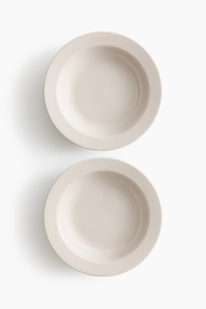 Deux bols beige clair sont présentés en vue aérienne, chacun avec un large rebord et un intérieur peu profond et incurvé. Ils sont alignés verticalement, l'un au-dessus de l'autre. - 1