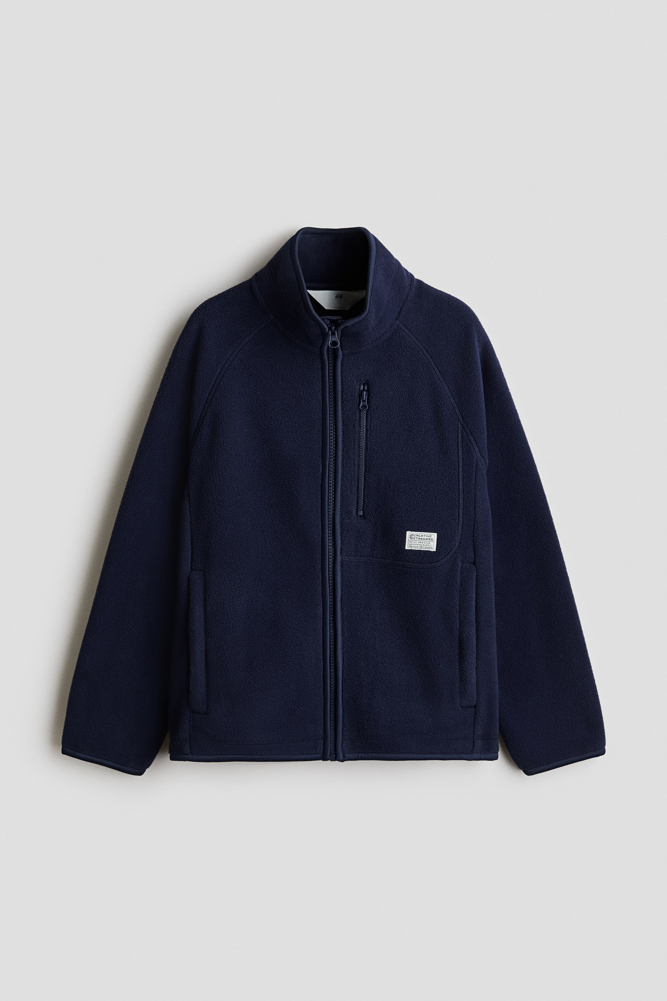 Se større billede: En marineblå fleecejakke vises forfra med en stående krave, fuld lynlås foran og lange raglanærmer. Den er designet med elastiske kanter ved ærmegab og kant samt en brystlomme med lynlås og to sidelommer med lynlås.