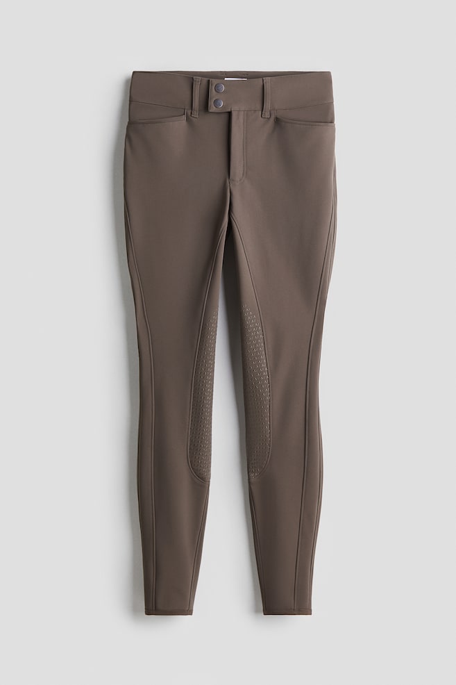 A pair of brown equestrian breeches is shown from the FRONT, featuring a double-button and zipper closure, small horizontal front pockets, and textured silicone grip panels on the inner legs. - 1