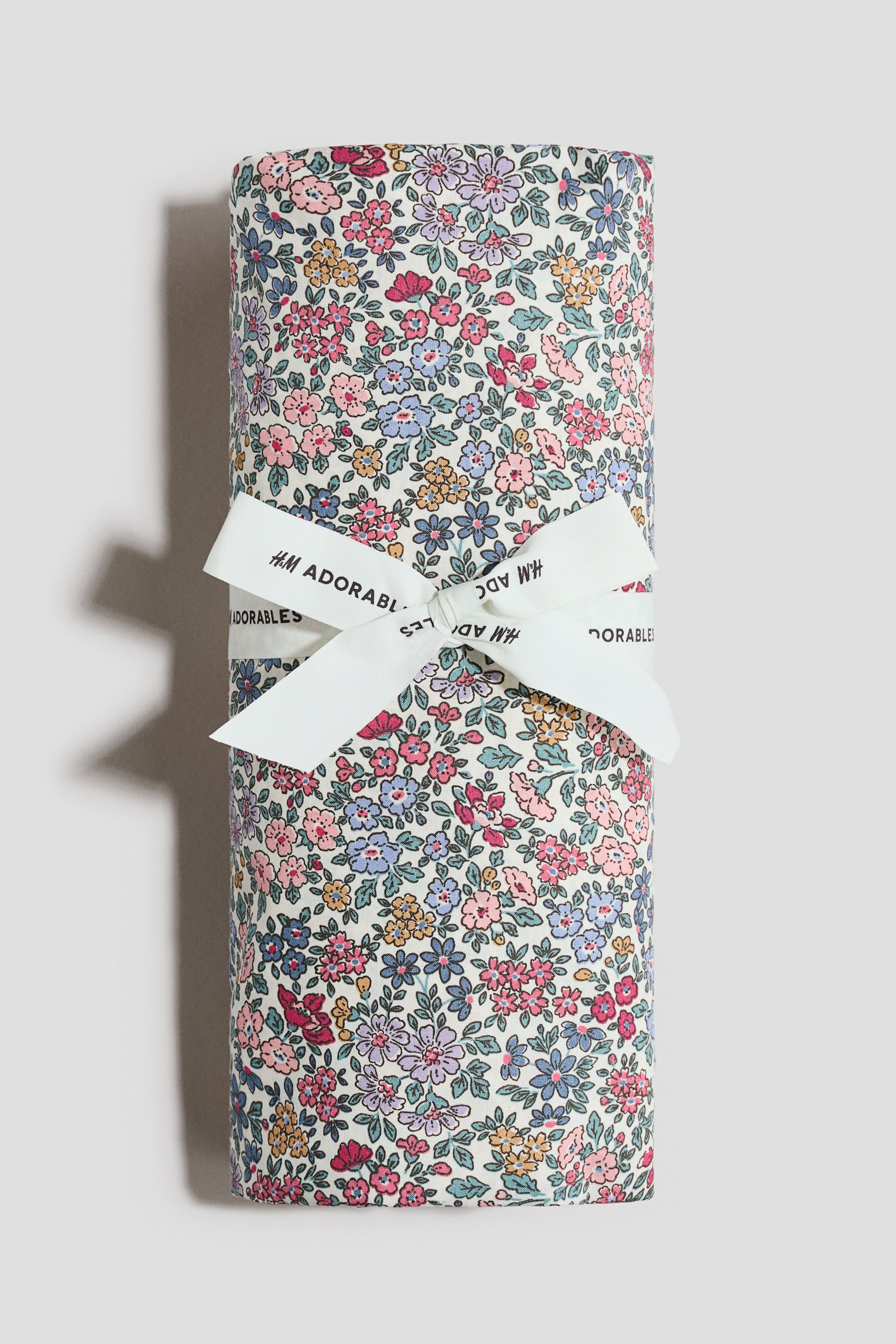 이미지 확대 보기: 장식이 있는 시트를 말아놓은 상태로 소개하고 있는데, 밝은 배경에 작은 분홍색, 파란색, 보라색, 노란색 꽃들이 전체적으로 인쇄되어 있습니다. "H.M ADORABLES" 텍스트가 있는 하얀 리본이 말아놓은 시트를 둘러싸고 리본 모양으로 묶여 있습니다.