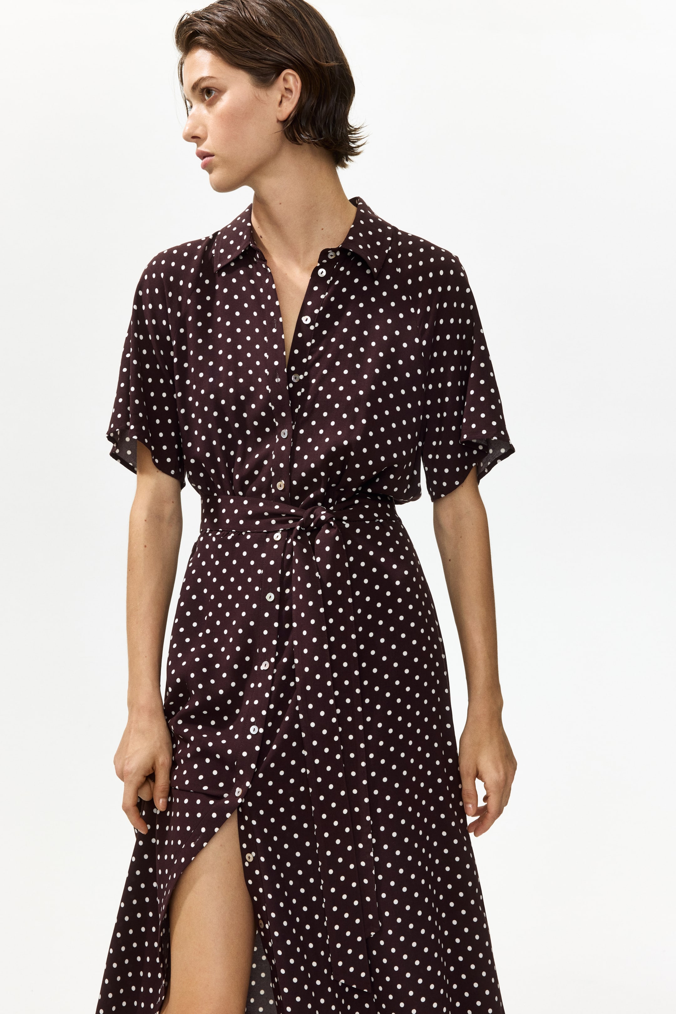 View larger image: A dark maroon shirtdress with white polka dots features a pointed collar, a partial button-front placket worn open at the top, and short, loose sleeves. A self-fabric tie belt cinches the waist, and the flowing skirt reveals a high left-side slit.