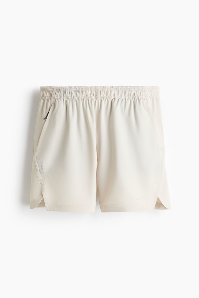 Pantalonii scurți sport sunt prezentați într-o culoare bej deschis, cu fața spre înainte, având o talie elasticizată și un buzunar cu fermoar pe partea stângă. Deschiderile laterale sunt prezente la terminațiile picioarelor, cu textul "DRYMOVE" poziționat vertical pe panoul lateral stâng și "MOVE" orizontal pe panoul frontal inferior drept. - 1