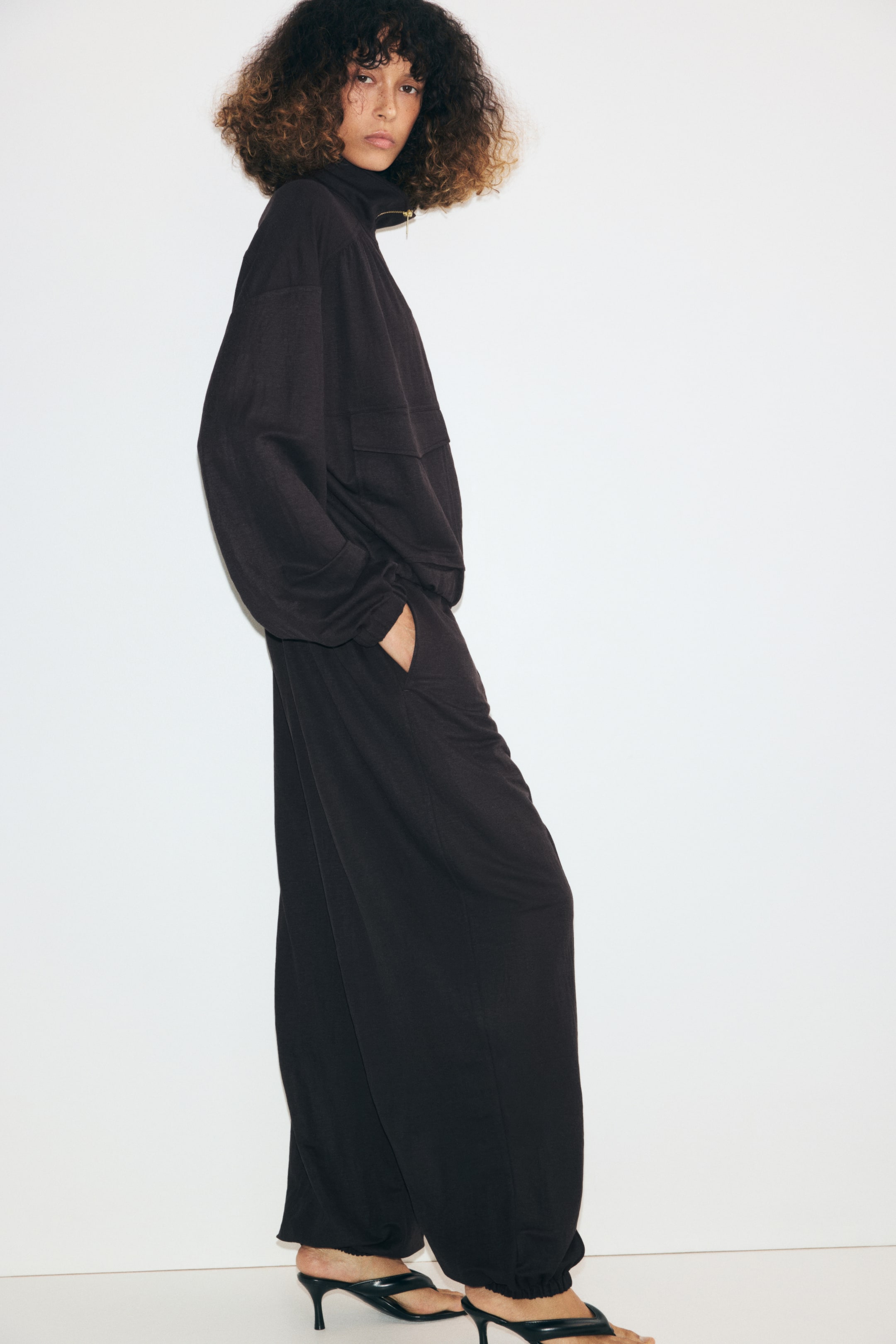 Ampliar la imagen: La modelo lleva puestos unos pantalones globo de jersey color café oscuro y corte holgado, mientras coloca una mano dentro del bolsillo lateral. Los pantalones, a la altura del tobillo, cuentan con dobladillos fruncidos con elástico y se combinan con una blusa a juego de cuello alto y sandalias negras de tacón.