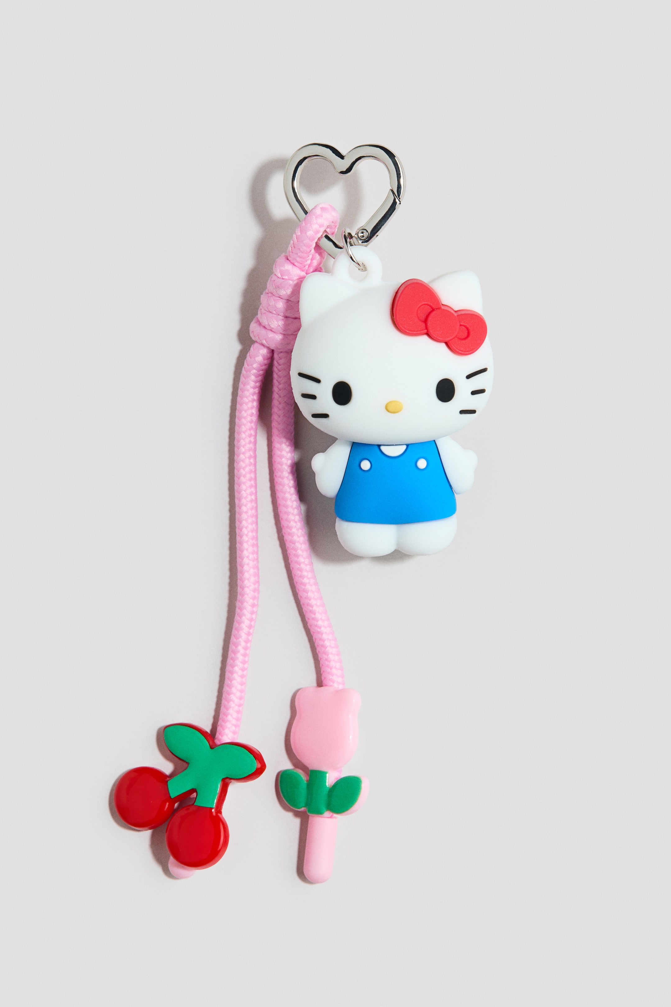 Grotere afbeelding bekijken: Een tasbedel met een zilverkleurige, hartvormige karabijnhaak aan de bovenkant, verbonden met een geknoopte roze gevlochten koord. Aan de koord hangt een witte Hello Kitty-figuur met een rode strik en blauwe jurk, een rode kersenbedel en een roze tulpenbedel, die allemaal hun vooraanzichten tonen.