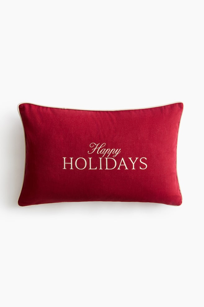 Un coussin décoratif rectangulaire rouge est présenté de face, avec les mots « Happy HOLIDAYS » brodés en lettres couleur crème au centre. Le coussin est fini avec un passepoil assorti de couleur crème le long des bords extérieurs. - 1