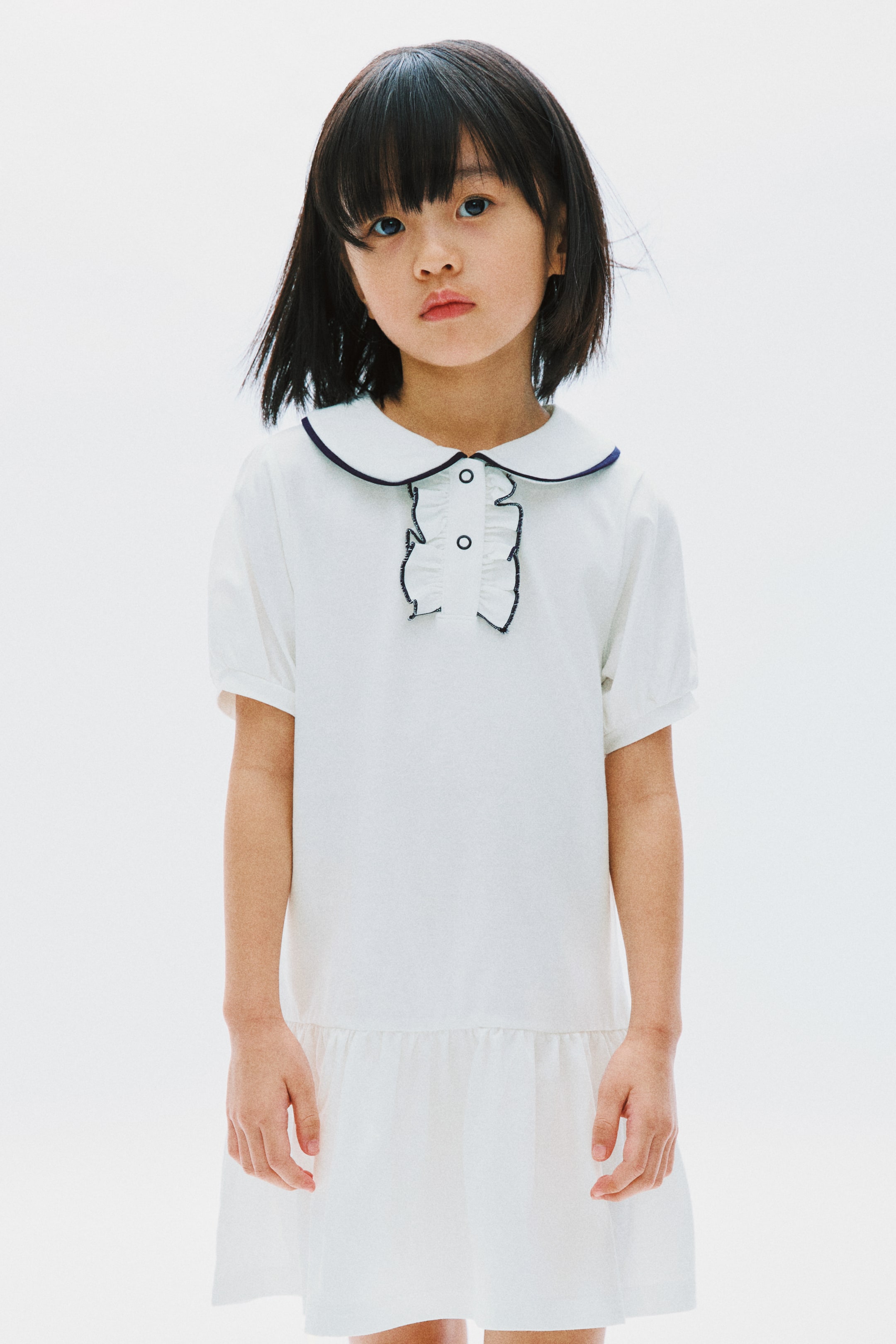 View larger image: This white dress showcases a rounded Peter Pan collar accented with a dark trim, short puffed sleeves, and a ruffled front placket adorned with two buttons. It features a gathered hemline, perfect for a chic and elegant look.