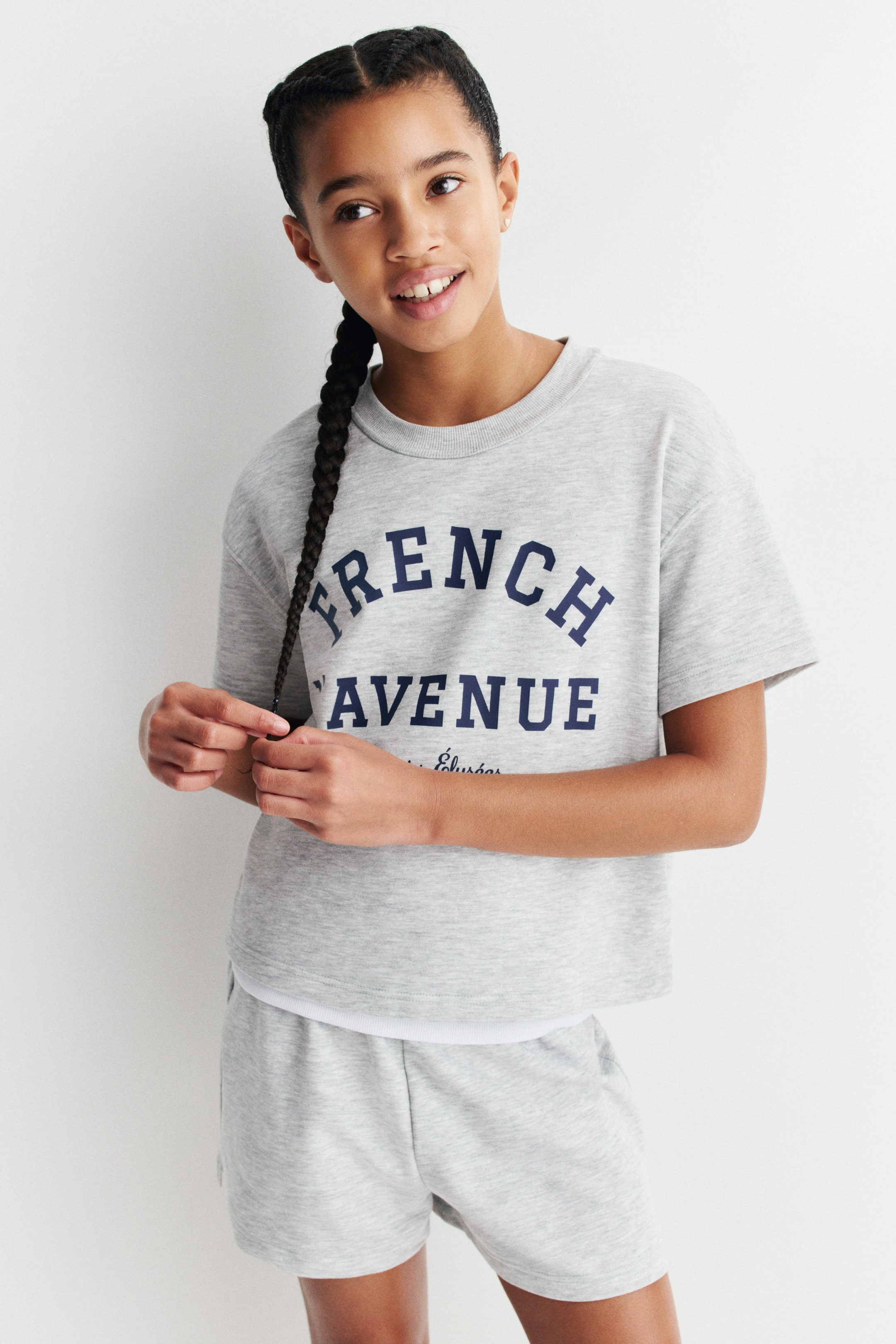 Grösseres Bild ansehen: Ein graues Marl-Kurzarm-Sweatshirt und passende Shorts sind nach vorne gerichtet. Das Sweatshirt hat einen gerippten Rundhalsausschnitt, überschnittene Schultern und "FRENCH AVENUE" in Dunkelblau auf der Vorderseite aufgedruckt, mit einer weißen Schicht, die unter dem verkürzten Saum hervorschaut, während die passenden Shorts eine lockere Passform aufweisen.