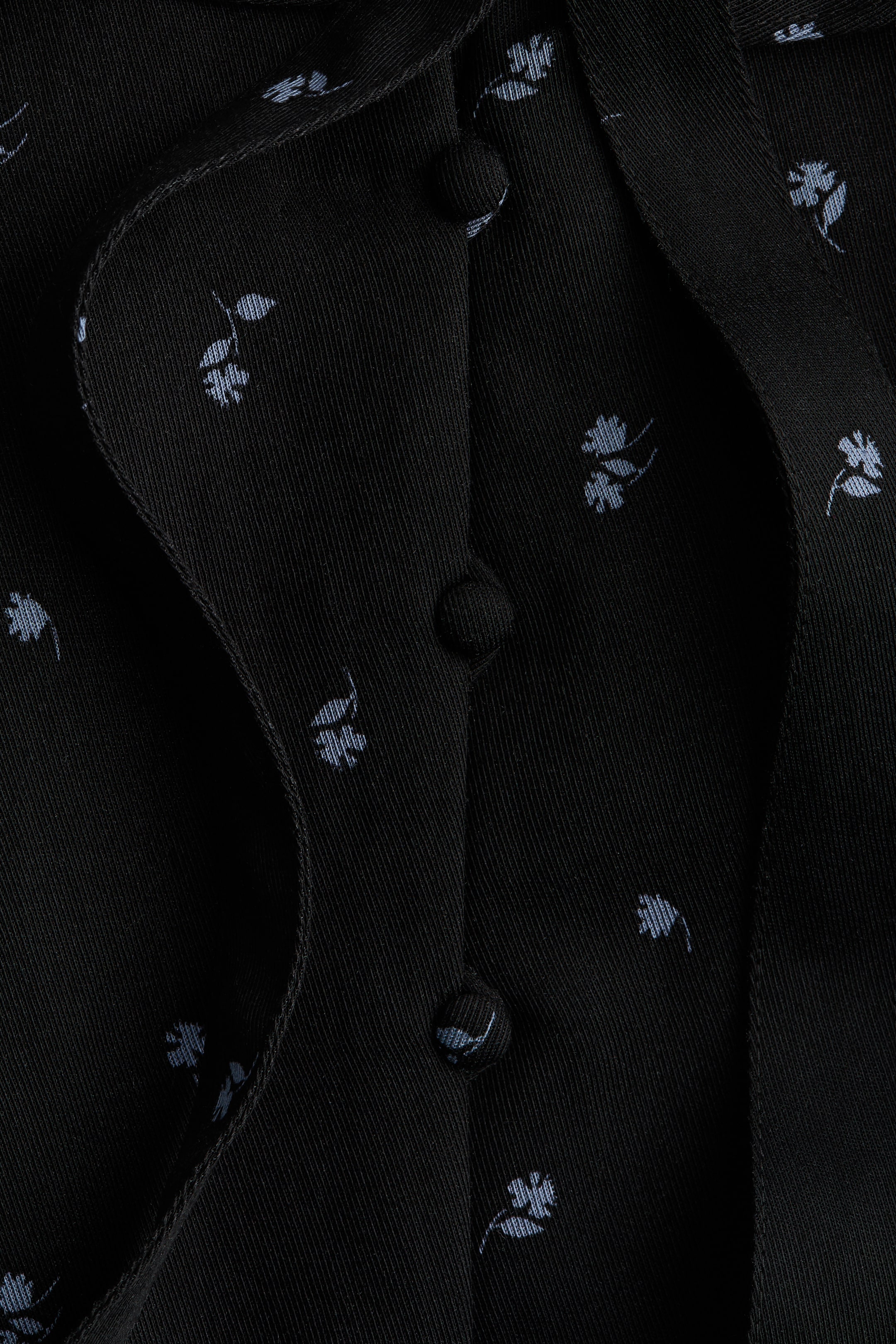 Xem ảnh lớn hơn: Một chiếc áo màu đen có in họa tiết hoa màu xanh nhạt tinh tế. Chiếc áo nổi bật với hàng nút với ba chiếc nút bọc vải ở phía TRƯỚC, nơi hai mảnh vải gặp nhau và trùng nhau.