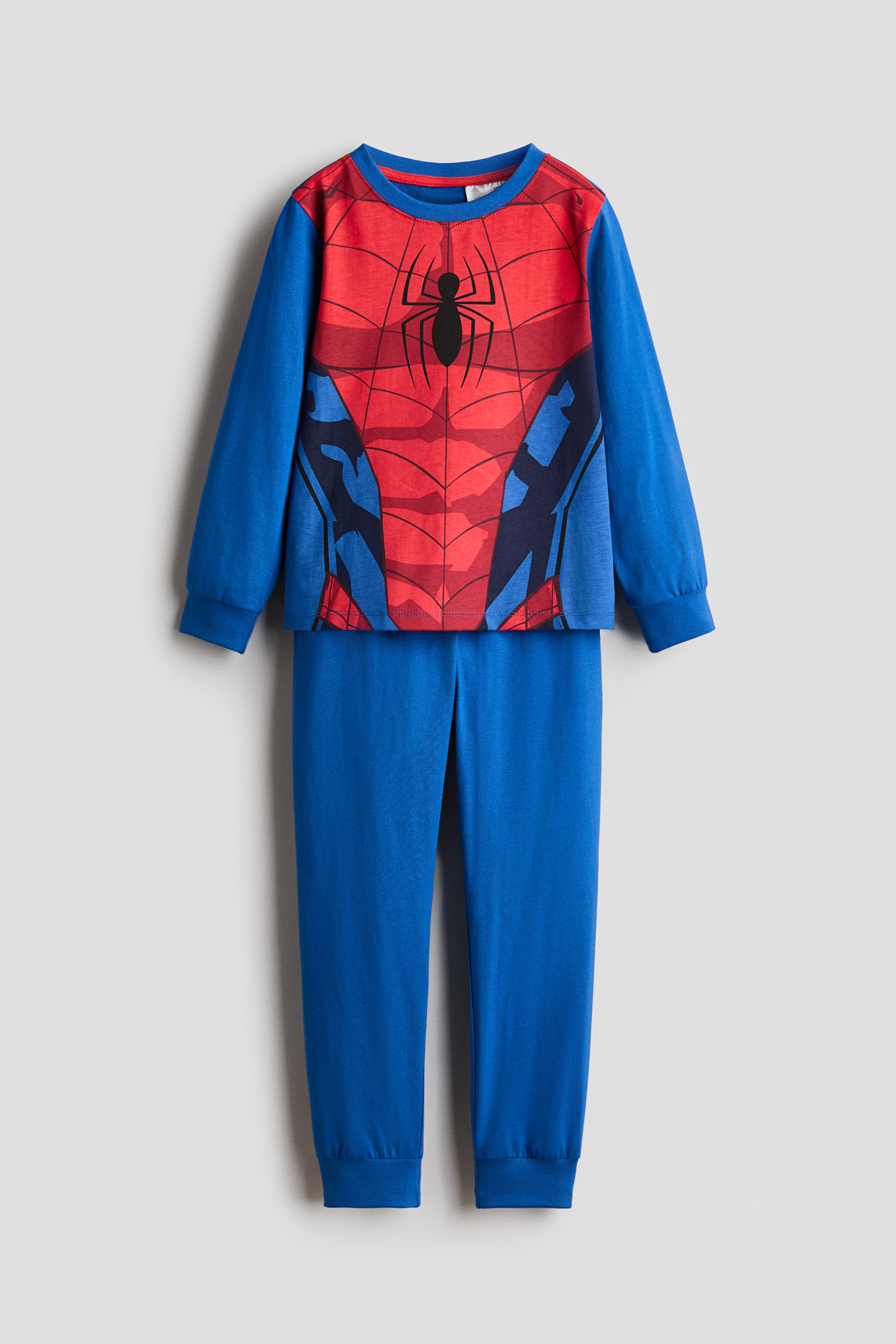 Vis større bilde: Et pyjamasett med en trykt topp og matchende underdel, presentert flatt fra forsiden. Toppen har et rødt og blått Spiderman-kostymedesign med en svart edderkopplogo på brystet, en rund ribbestrikket hals og lange ermer med ribbestrikkede mansjetter. De blå underdelene har en elastisk midje og ribbestrikkede kanter.