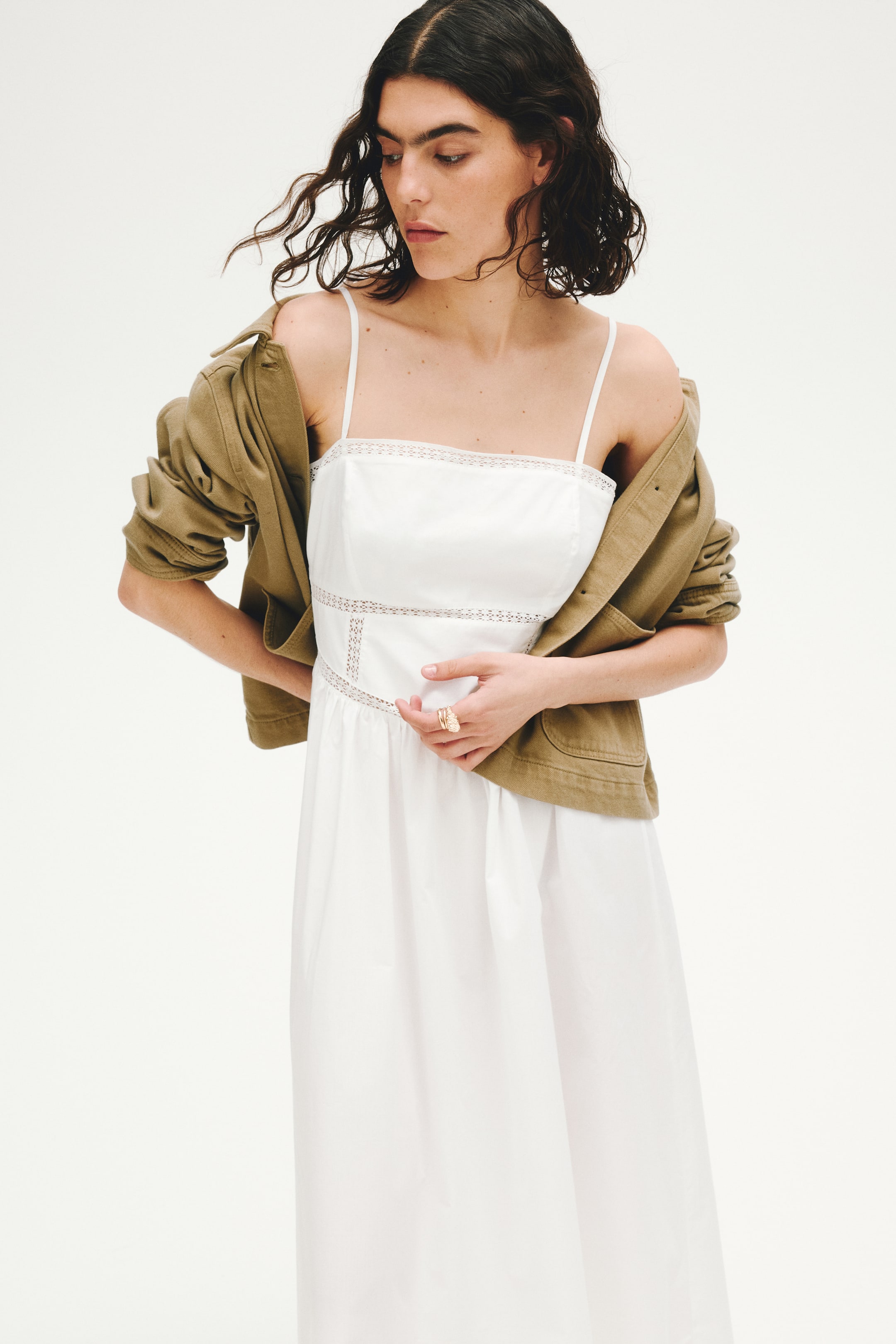 View larger image: This white dress showcases thin spaghetti straps and a square neckline adorned with delicate lace trim. A horizontal band of eyelet lace defines the waist, sitting above a gathered, flowing skirt.
