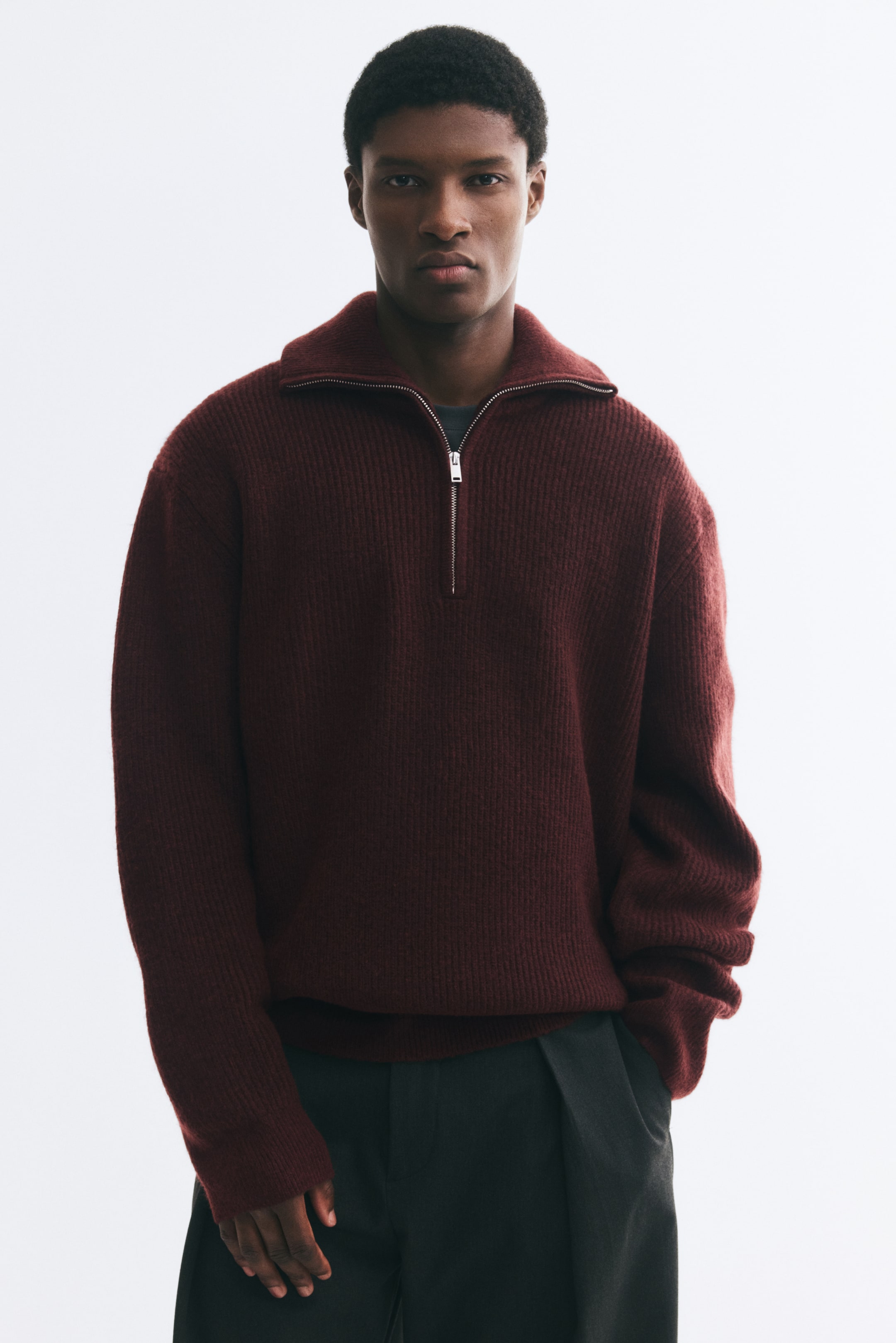 View larger image: A burgundy rib-knit sweater is being worn by the model, featuring a relaxed fit, a partially unzipped stand collar, and long sleeves. The sweater is layered over a dark undershirt and paired with dark trousers.