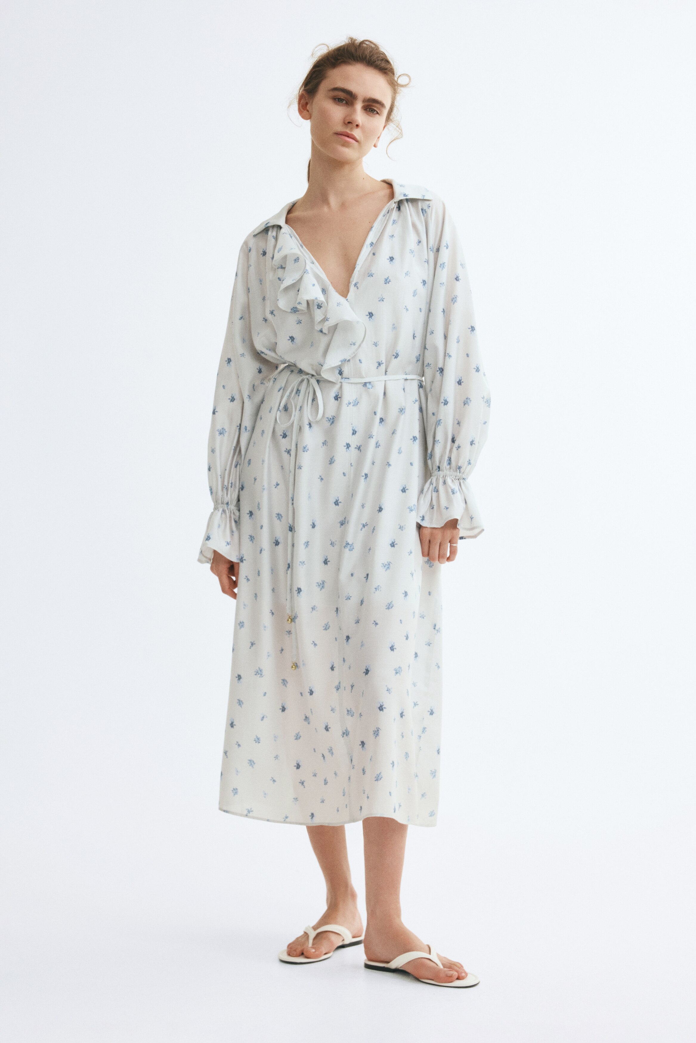 Facing front, this light grey midi dress features a scattered blue floral pattern and a pointed collar with a vertical ruffled placket. The dress has long balloon sleeves with elasticated ruffled cuffs and a thin drawstring tie cinching the waist.