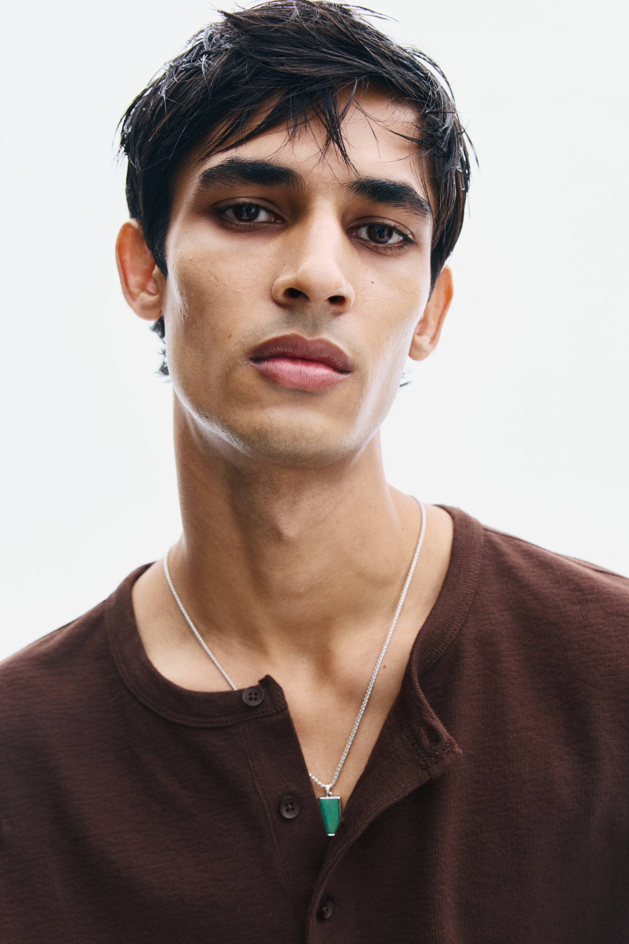 View larger image: A silver chain necklace featuring a small, rectangular green stone pendant is being worn by the model. The necklace is resting against a dark brown, textured henley shirt with button details.