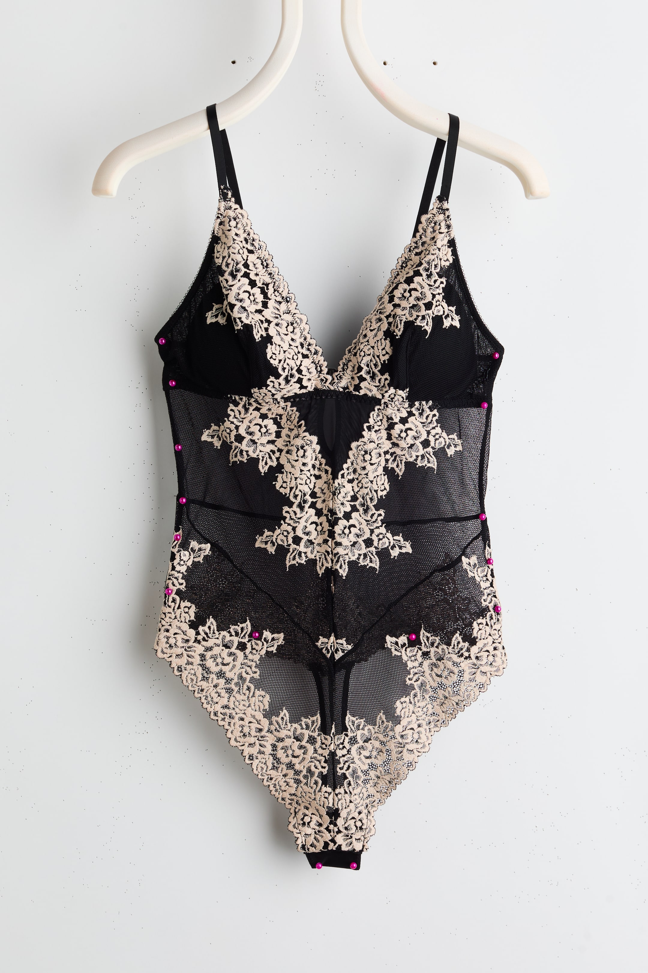View larger image: A black bodysuit featuring intricate beige floral lace detailing over black mesh, forming triangle cups and extending across the torso. It is designed with thin adjustable shoulder straps, and small magenta beads accent the lace.
