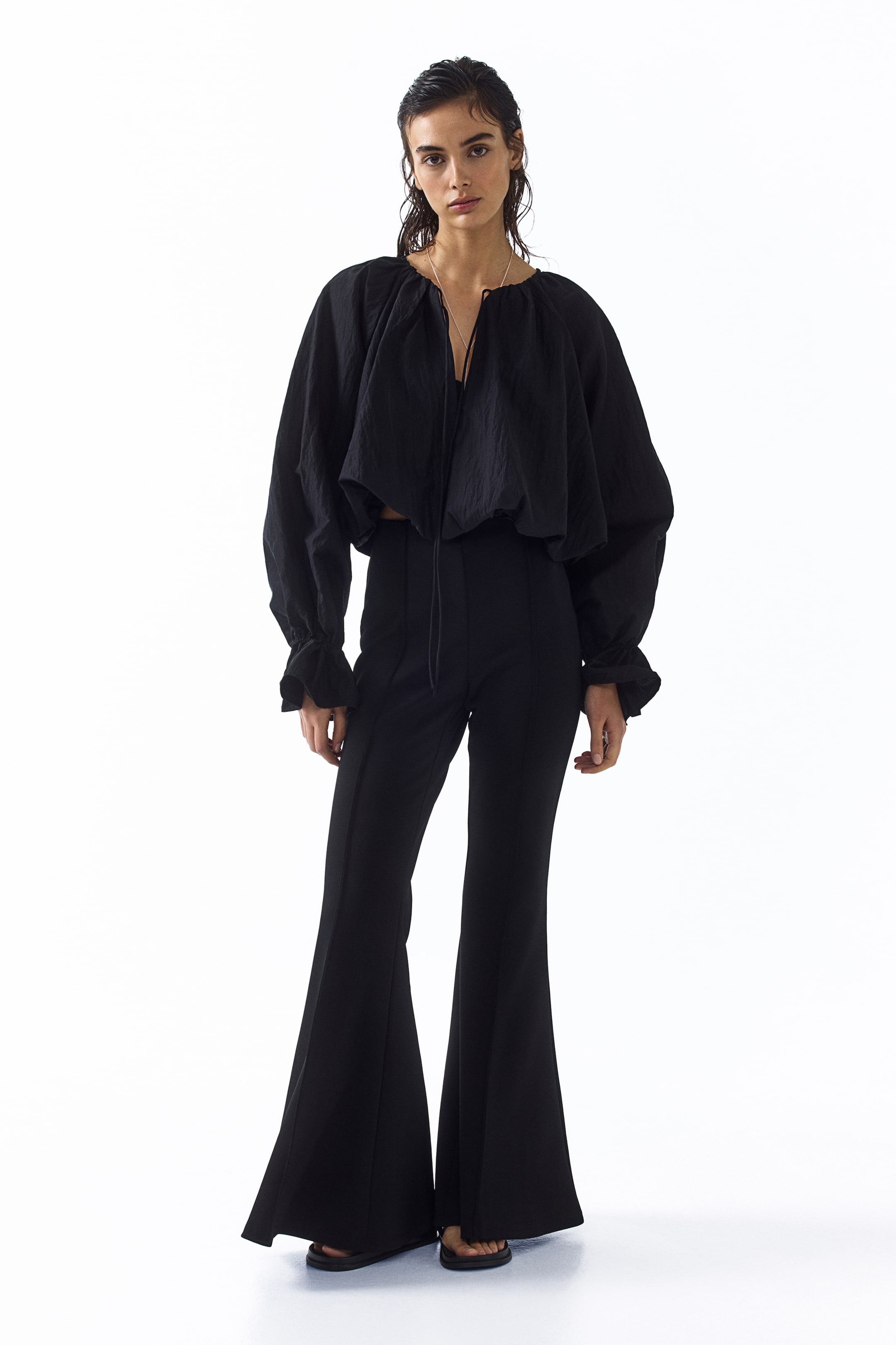 Ver imagen más grande: Un conjunto negro de dos piezas presenta una blusa corta y voluminosa con un escote fruncido con cordón, una textura arrugada y puños con volantes, sobre un camisón oscuro. Los pantalones de cintura alta a juego presentan una silueta acampanada y costuras verticales en la parte delantera.