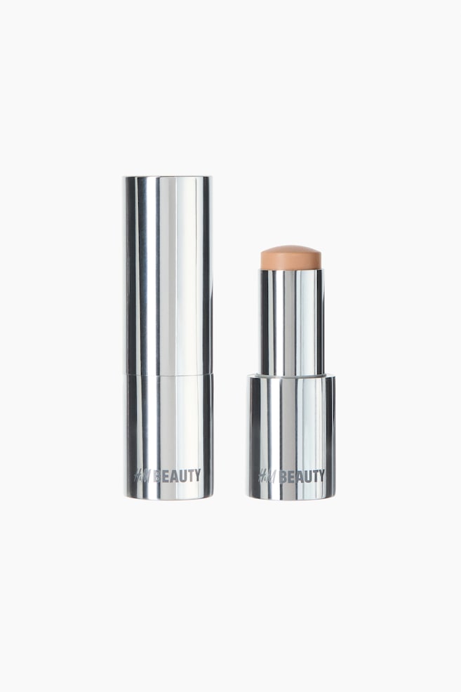 Two cylindrical beauty sticks are standing upright. The stick on the left is closed, featuring a metallic silver casing displaying an 'H&M BEAUTY' logo near its base. The stick on the right is open, revealing a light beige/tan-colored product extending from its metallic silver base, also displaying the 'H&M BEAUTY' logo. - 1