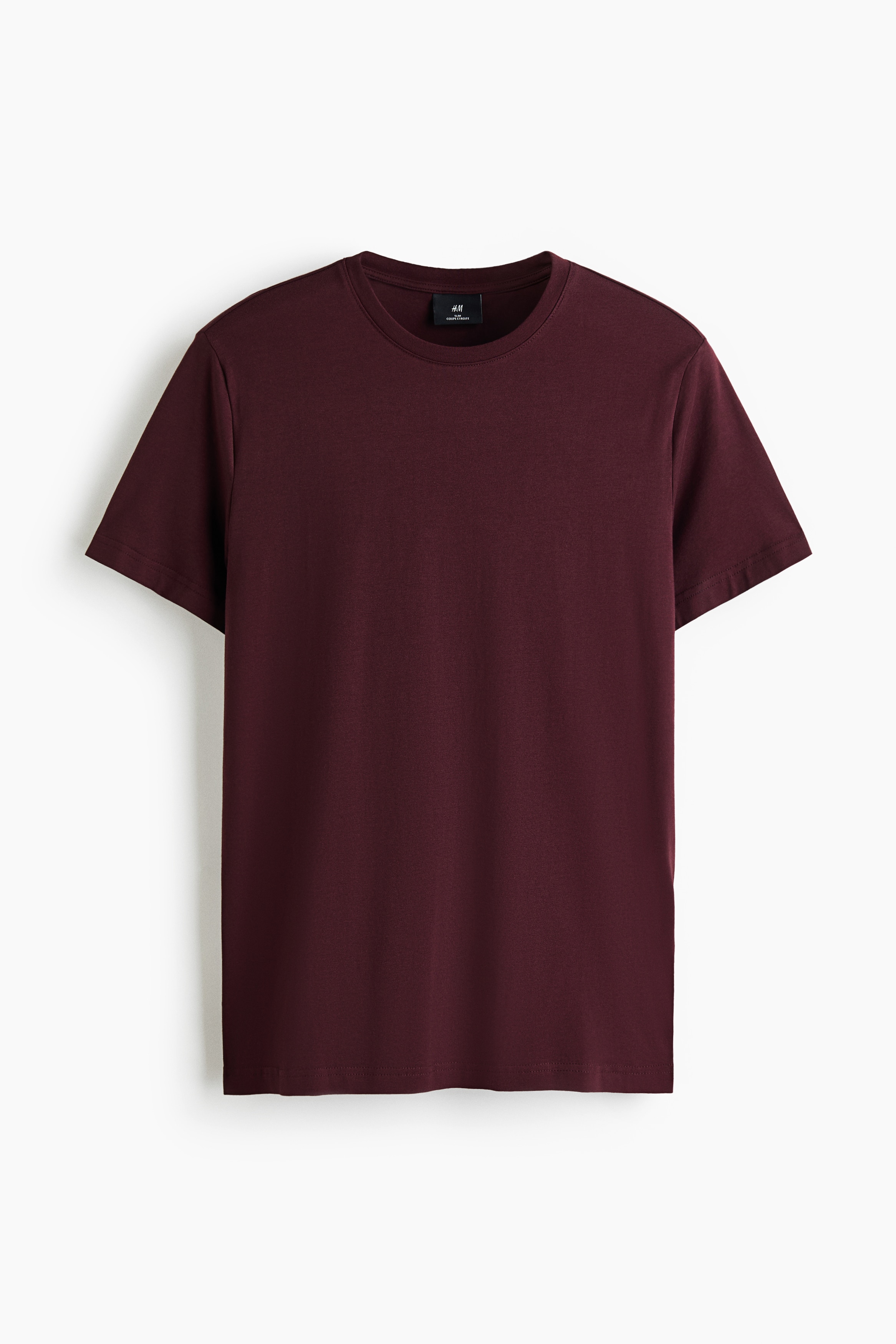 Una t-shirt slim fit bordeaux in jersey di cotone leggero è indossata dalla modella, con scollo rotondo rifinito a costine e maniche corte. La t-shirt è abbinata a jeans blu, con la modella che tiene entrambe le mani nelle tasche anteriori.