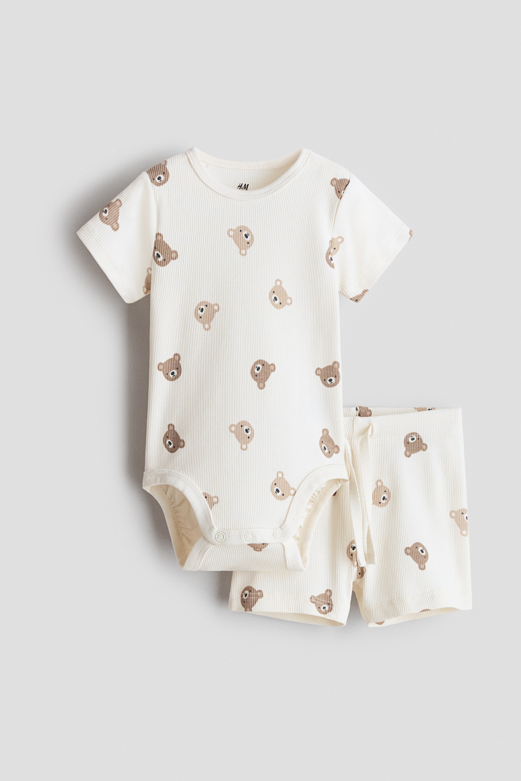 View larger image: A cream-coloured, ribbed jersey set features a short-sleeved bodysuit and matching shorts, both with an all-over brown bear head pattern on the front. The bodysuit has a round neckline and press-studs at the crotch, while the shorts have an elasticated waist with a visible drawstring.