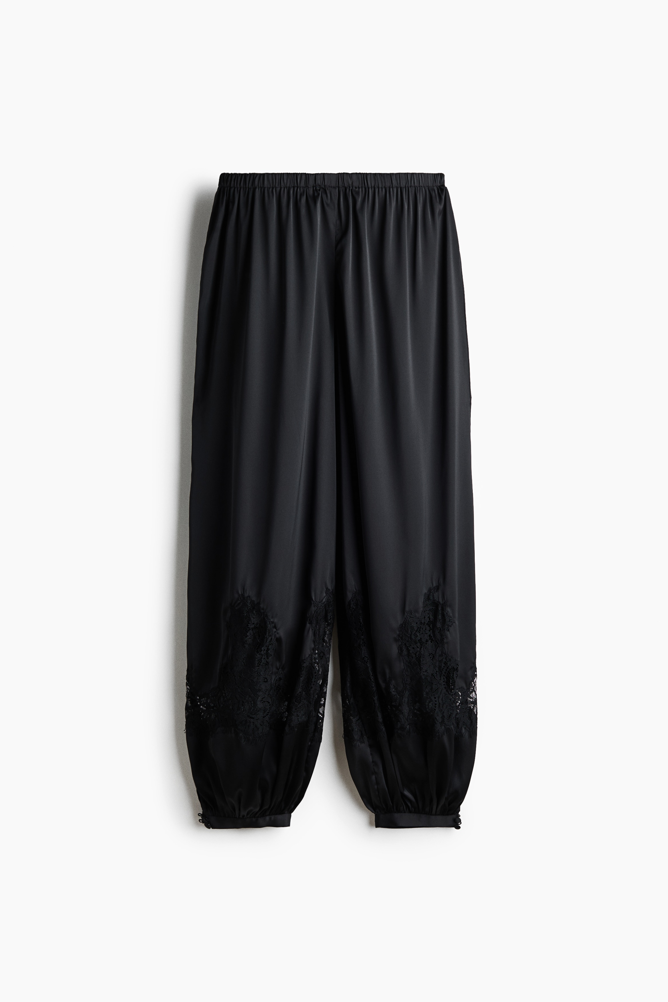 Black loose-fit satin pants with an elasticated waist, balloon legs, and sheer lace inserts near the ankles are worn by the model, who is standing in profile. The pants are styled with a matching black satin halter top and black platform slide sandals.