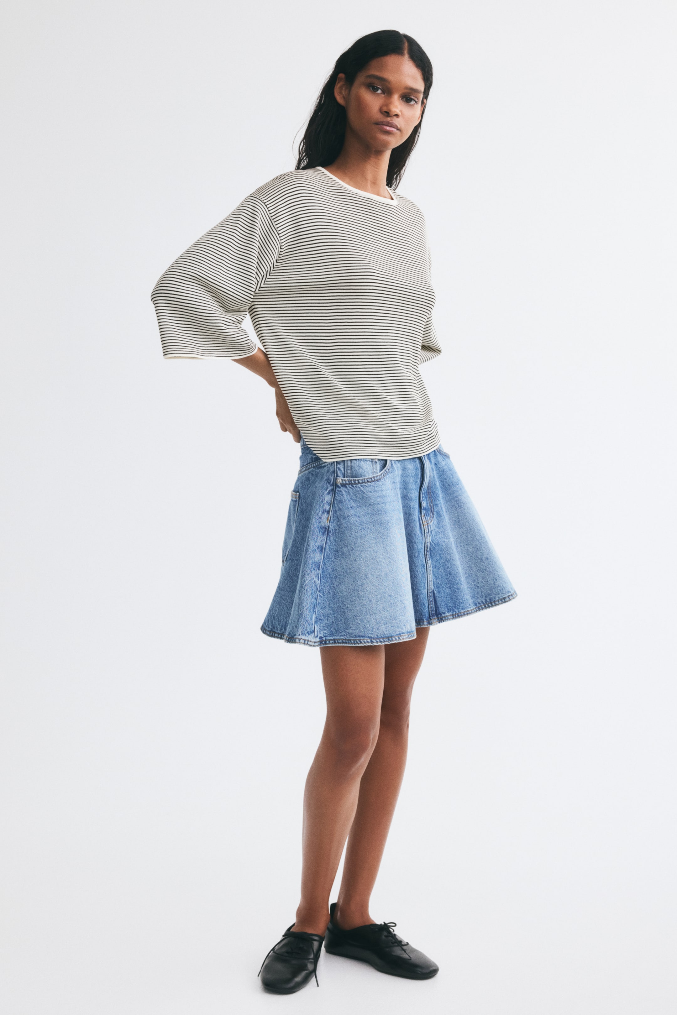 View larger image: A loose-fit, fine-knit striped sweater featuring a round neckline with a narrow trim, dropped shoulders, and 3/4-length sleeves is being worn by the model. The model is pairing the sweater with a light blue denim skirt and is placing her hands on her hips.