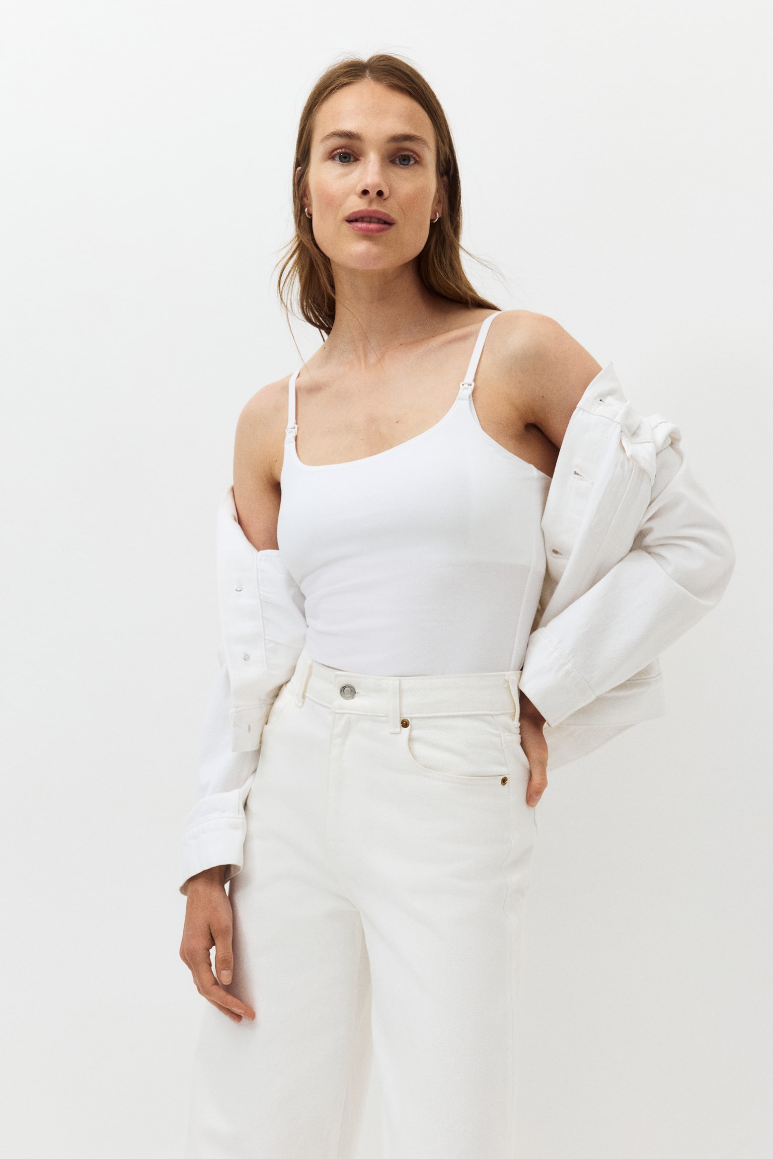 Presented from a front-right angle, this white camisole features slim shoulder straps, with a visible clip on one strap, and a slightly scooped neckline. It showcases a fitted silhouette, neatly tucked into white jeans. - 1
