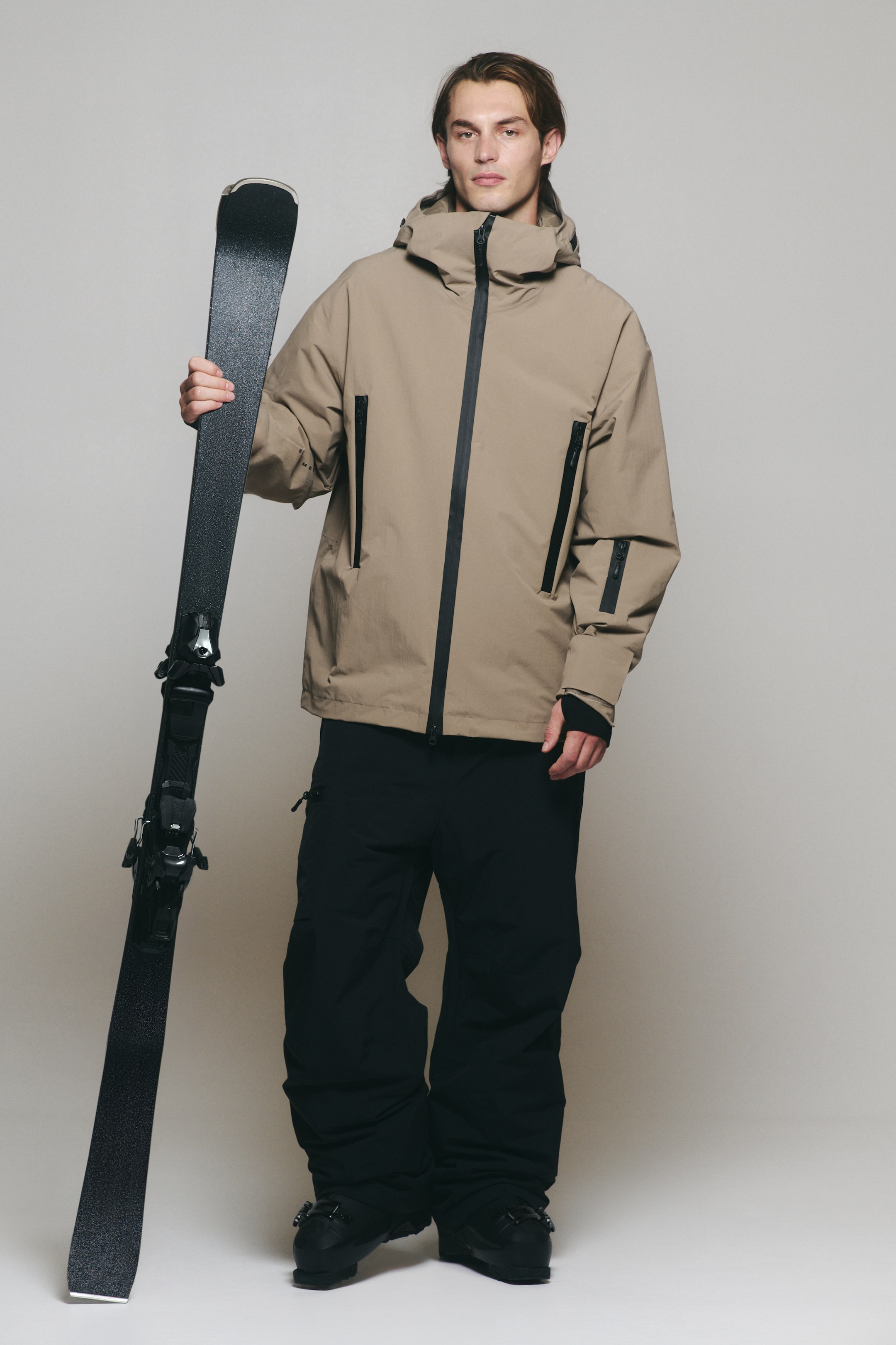 Agrandir l'image: Une veste de ski à capuche beige est portée par le modèle, assortie à un pantalon de ski noir ample et à des bottes de ski noires. Le modèle tient un seul ski noir à la verticale sur le côté.