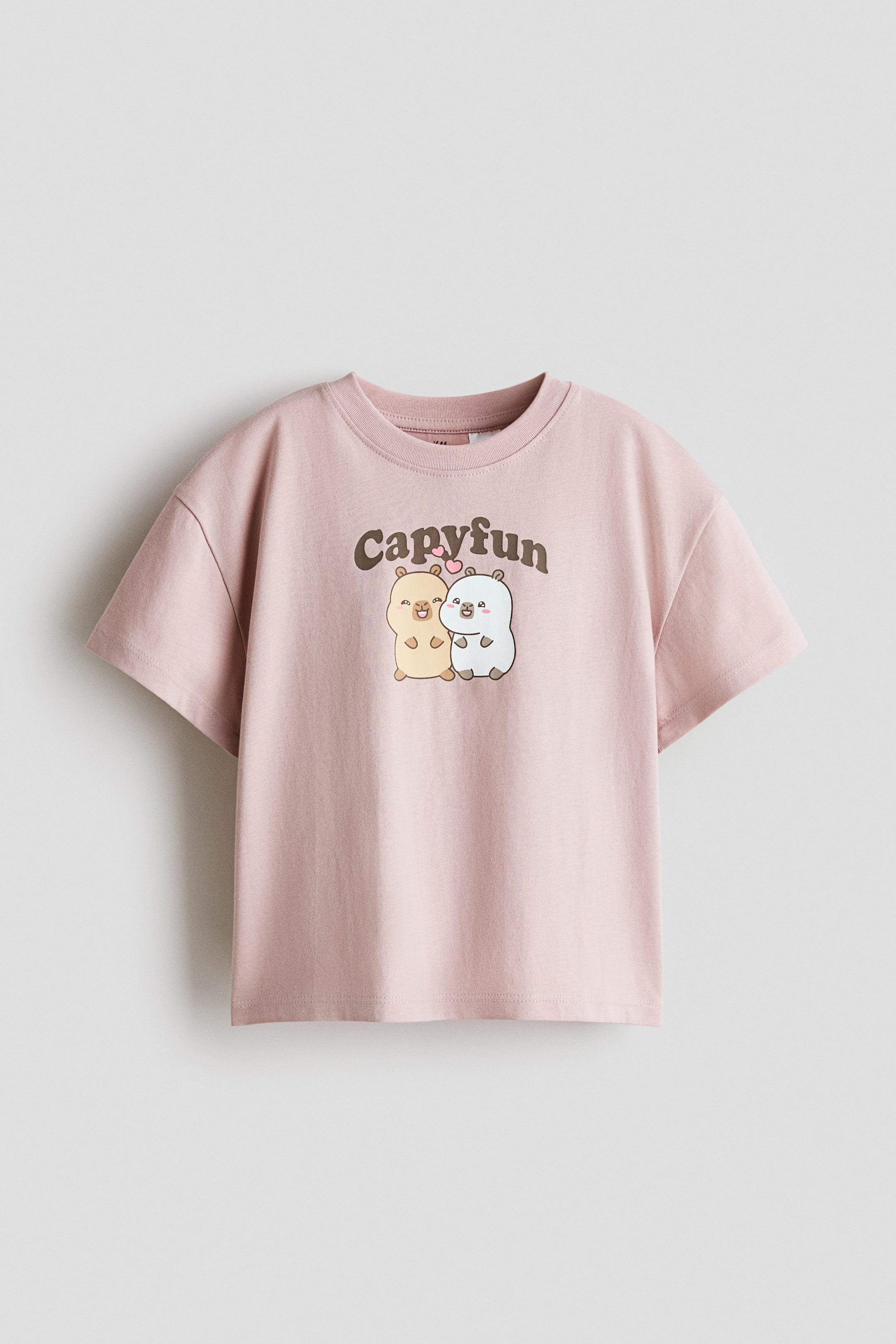 Xem ảnh lớn hơn: Một chiếc áo thun cotton jersey màu hồng được chụp chính diện, có cổ tròn, vai xuôi và tay ngắn. Mặt trước in hình đồ họa hai chú capybara hoạt hình bên dưới dòng chữ nâu uốn cong “Capyfun.”