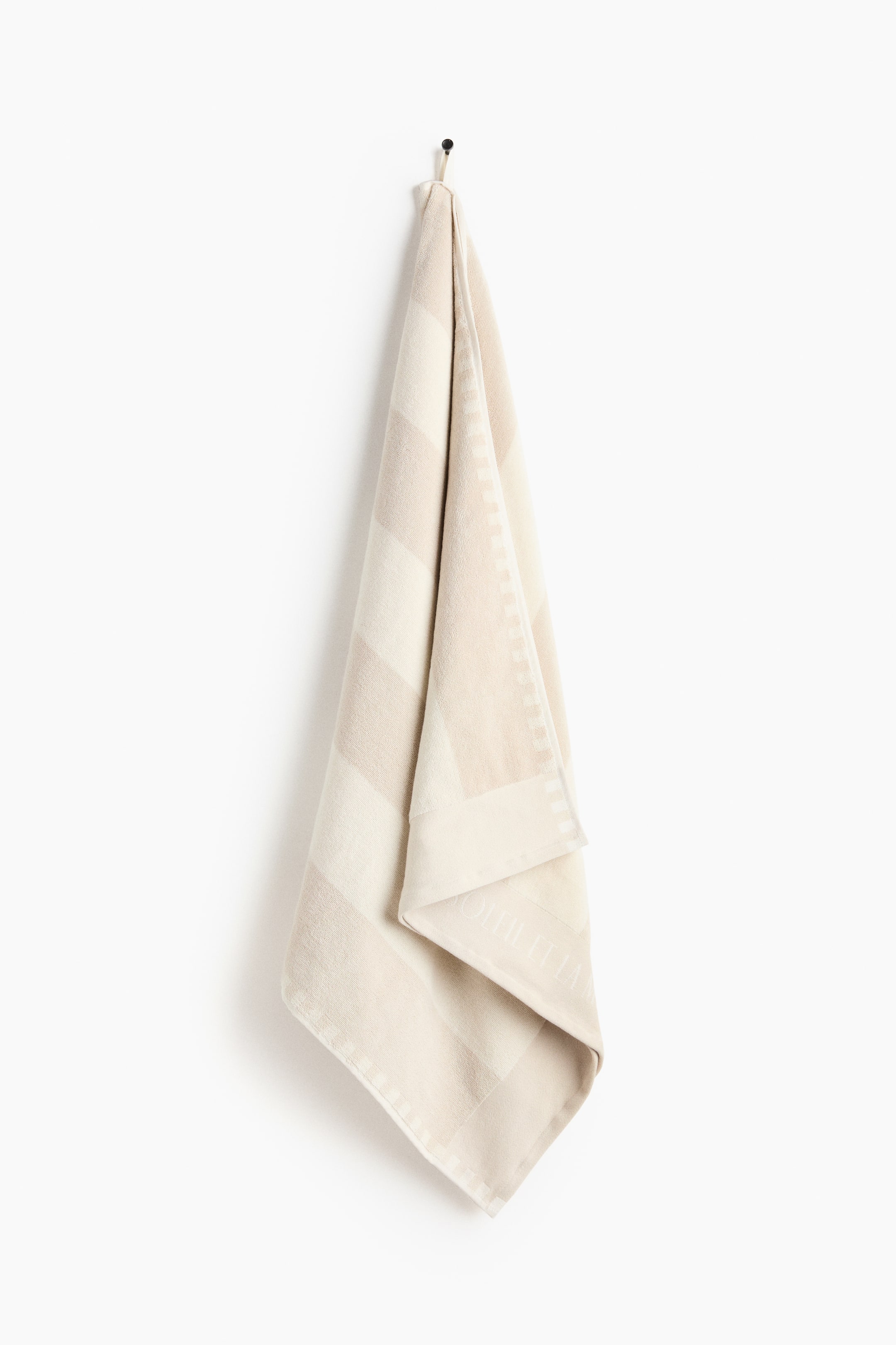 View larger image: A towel is showcasing broad alternating beige and cream diagonal stripes. It hangs vertically with gentle folds, revealing a patterned border and subtle text woven into the lower section.