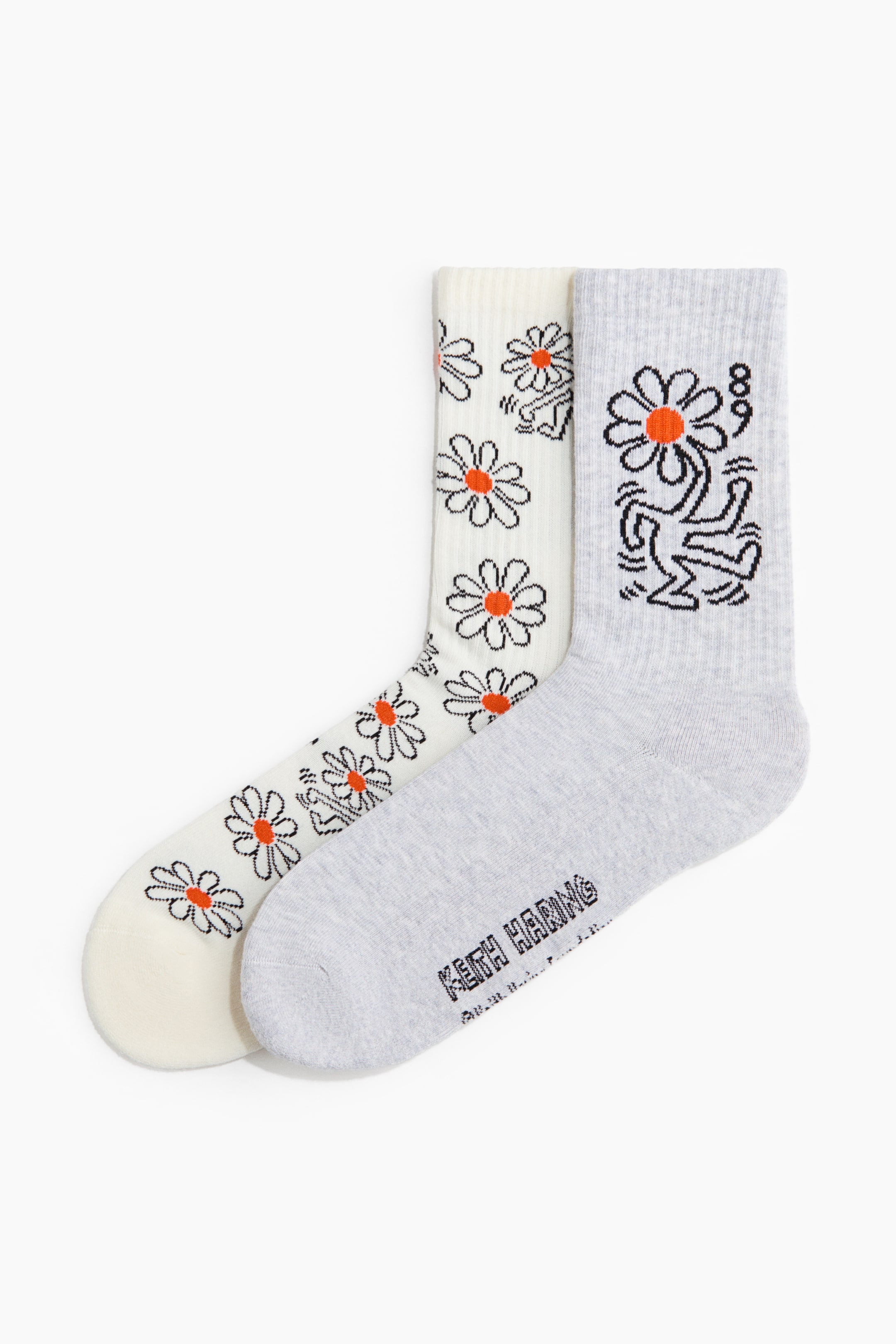 View larger image: Two socks are laid flat, slightly angled, each featuring a high, ribbed shaft. The off-white sock displays an all-over pattern of daisy motifs with orange centres and black outlines, interspersed with small black figures, while the light grey sock showcases a larger central motif of a black outlined figure holding a daisy with an orange centre, additional small black figures, and "KEITH HARING" text visible on the sole.