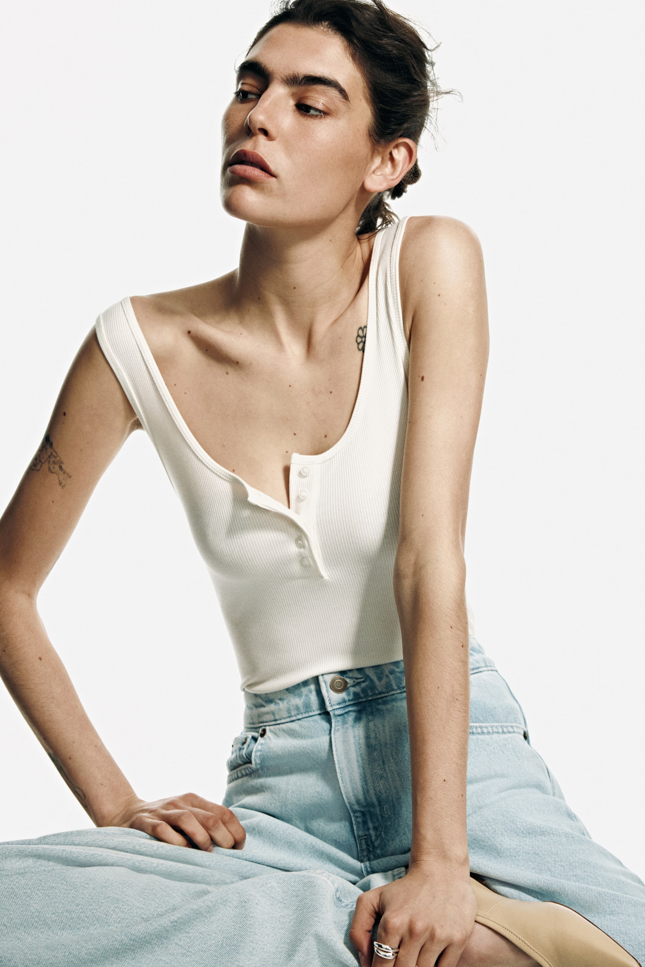 View larger image: The model is wearing a sleeveless body made from ribbed jersey, showcasing a scoop neckline, a front button placket, and narrow binding around the neckline and armholes. The model is posing with the body tucked into light-wash denim jeans.