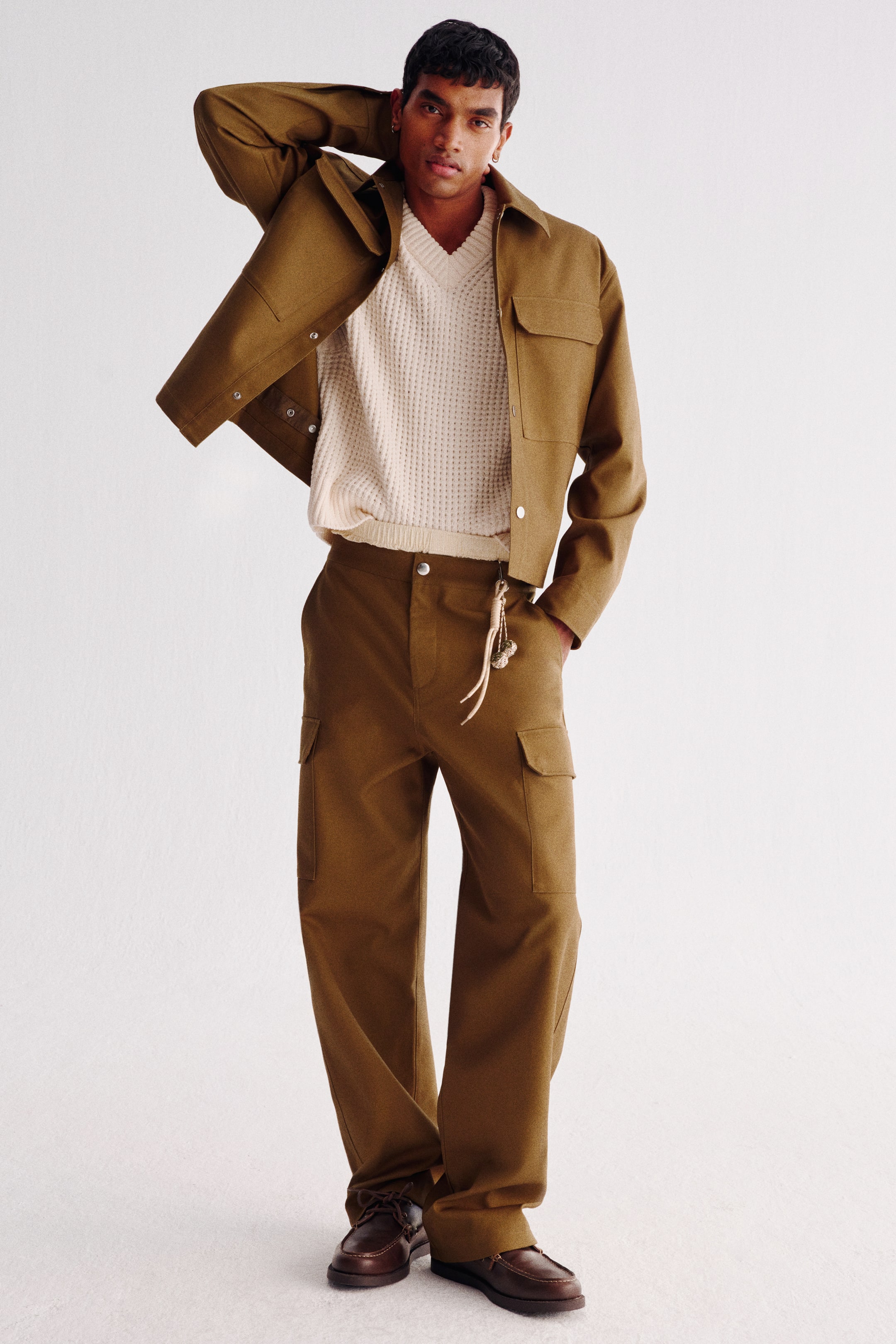View larger image: An olive green cropped jacket featuring two flap chest pockets is layered over an off-white V-neck waffle knit sweater. This is being paired with matching olive green cargo pants, which have side cargo pockets and a decorative charm hanging from a belt loop, and dark brown lace-up loafers.