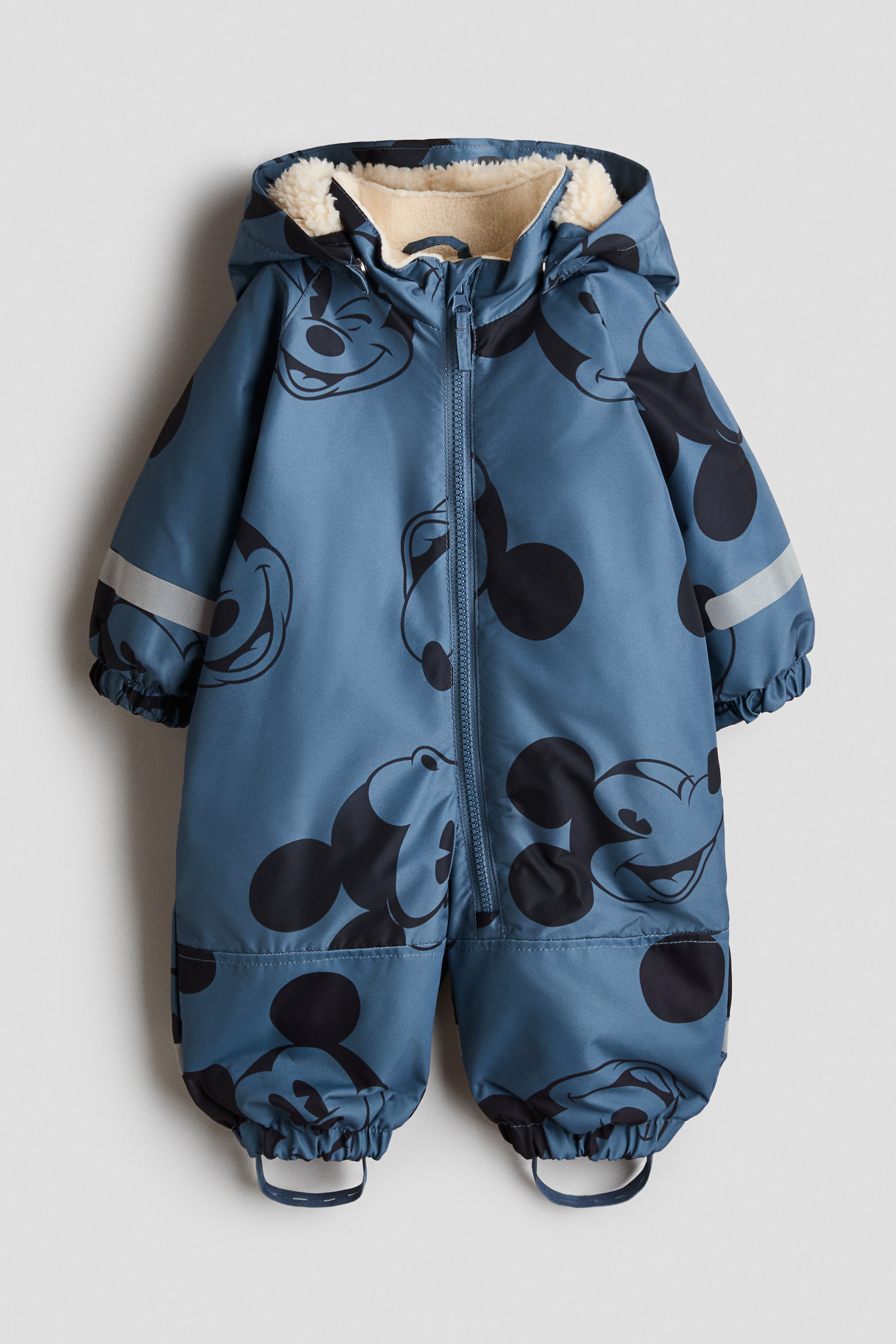 Visa större bild: En blå overall för barn visar ett upprepat mönster av svarta Mickey Mouse-ansikten, sett framifrån. Den har en krämfärgad huva fodrad med sherpa, en dragkedja framtill i mitten, resår vid ärmsluten med reflekterande ränder på ärmarna samt elastiska benslut med fotbyglar.