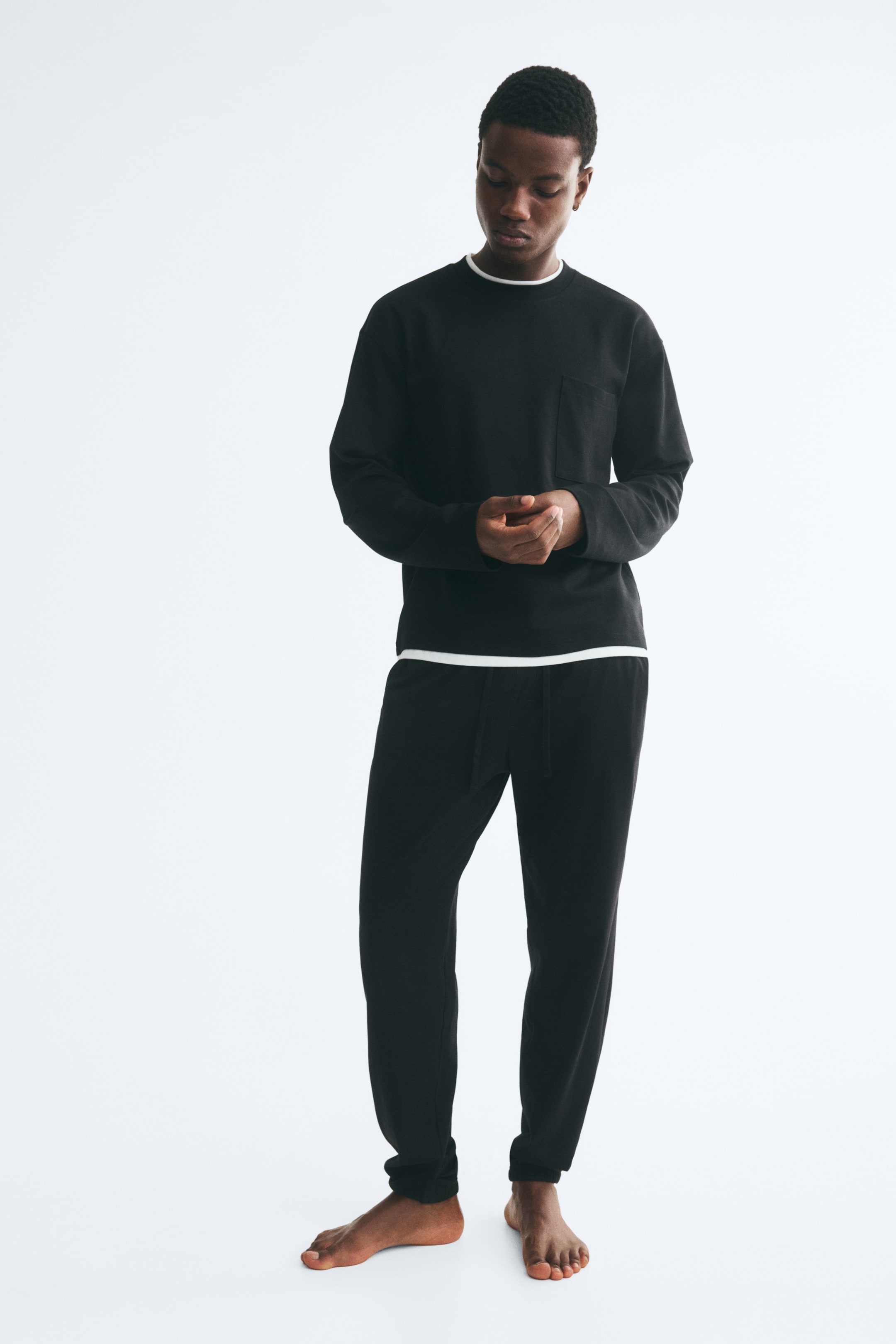 View larger image: Black cotton jersey track pants featuring a regular fit, an elasticated drawstring waist and ribbed hems are being worn by the model. The trousers are paired with a matching black long-sleeved top featuring white trim at the collar and hem.