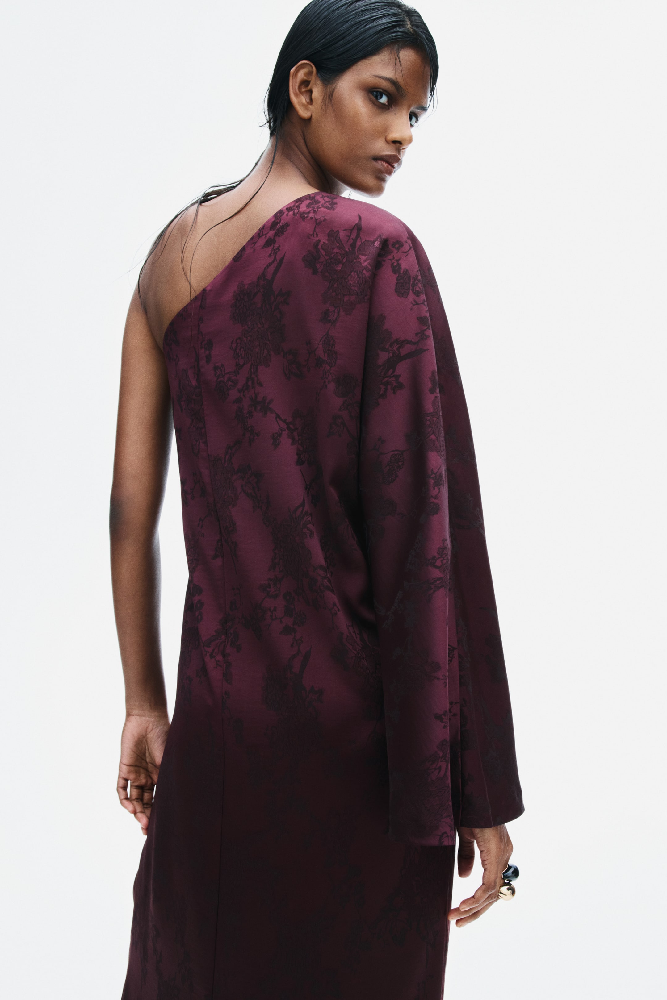 View larger image: A burgundy jacquard-weave satin dress with a floral pattern is being worn by the model, showcasing an asymmetrical one-shoulder cut with a single long, wide sleeve. The loose-fitting garment is displayed from behind as the model turns to look towards the camera.
