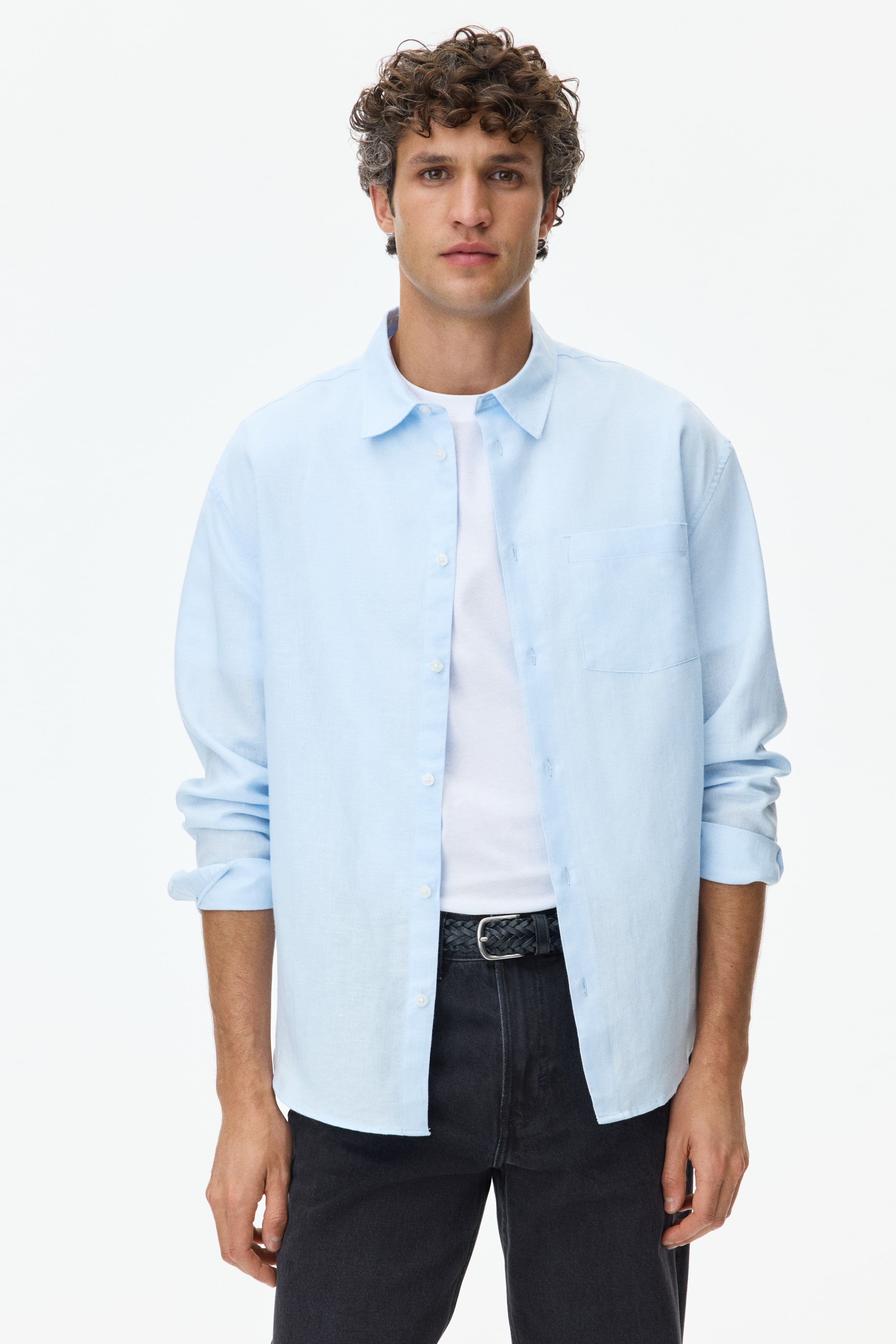 View larger image: A light blue long-sleeve shirt is being showcased front-facing, featuring a spread collar, a full button-down placket with white buttons, and a single patch pocket on the left chest. Its long sleeves are rolled up, and it is being worn open over a white t-shirt, displaying a relaxed fit.