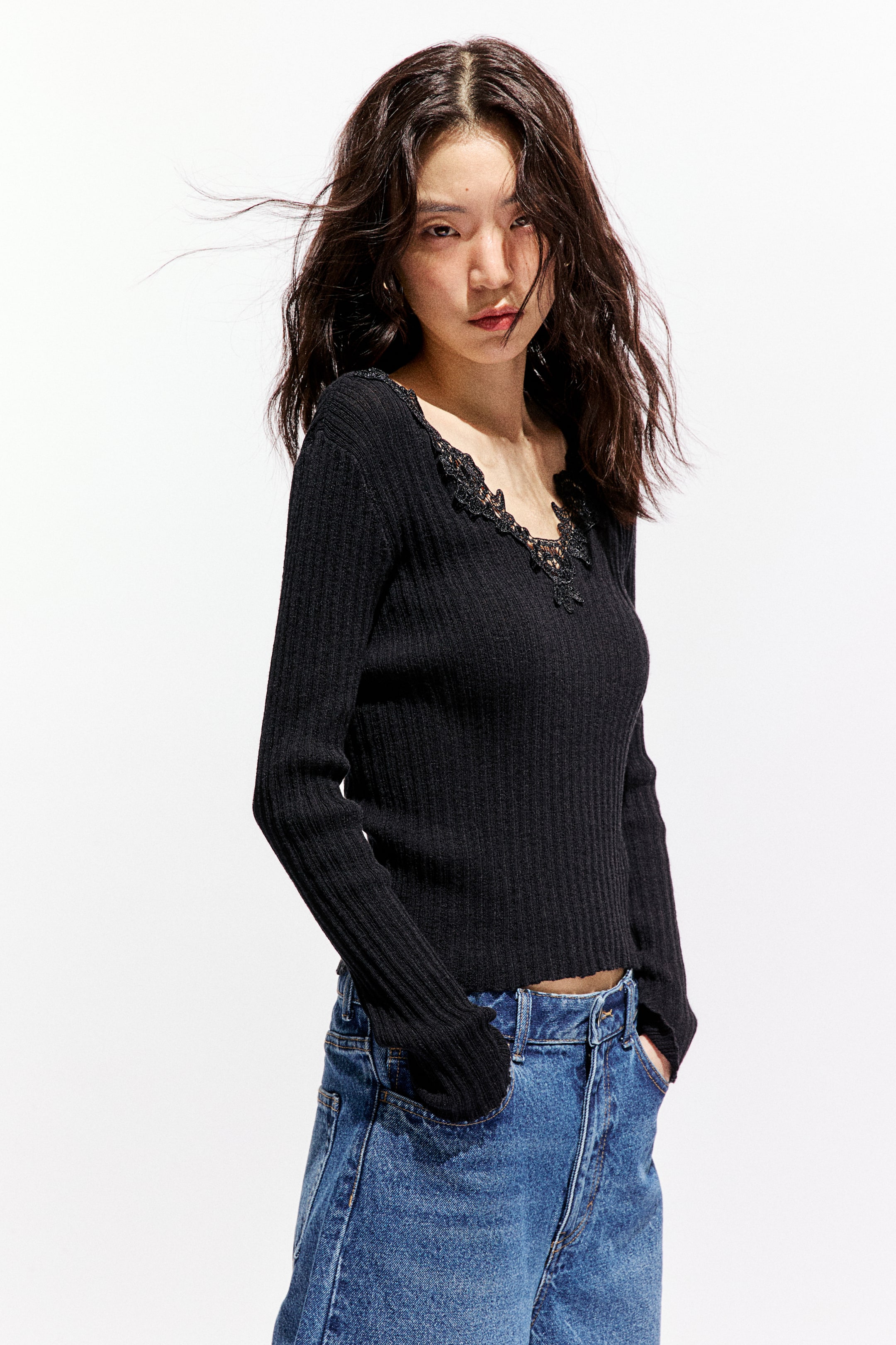拡大画像を表示: モデルが着用しているのは、レーストリム付きのVネックラインと長袖が特徴のフィットしたブラックリブニットトップです。彼女はブルージーンズのポケットに手を入れて立っています。