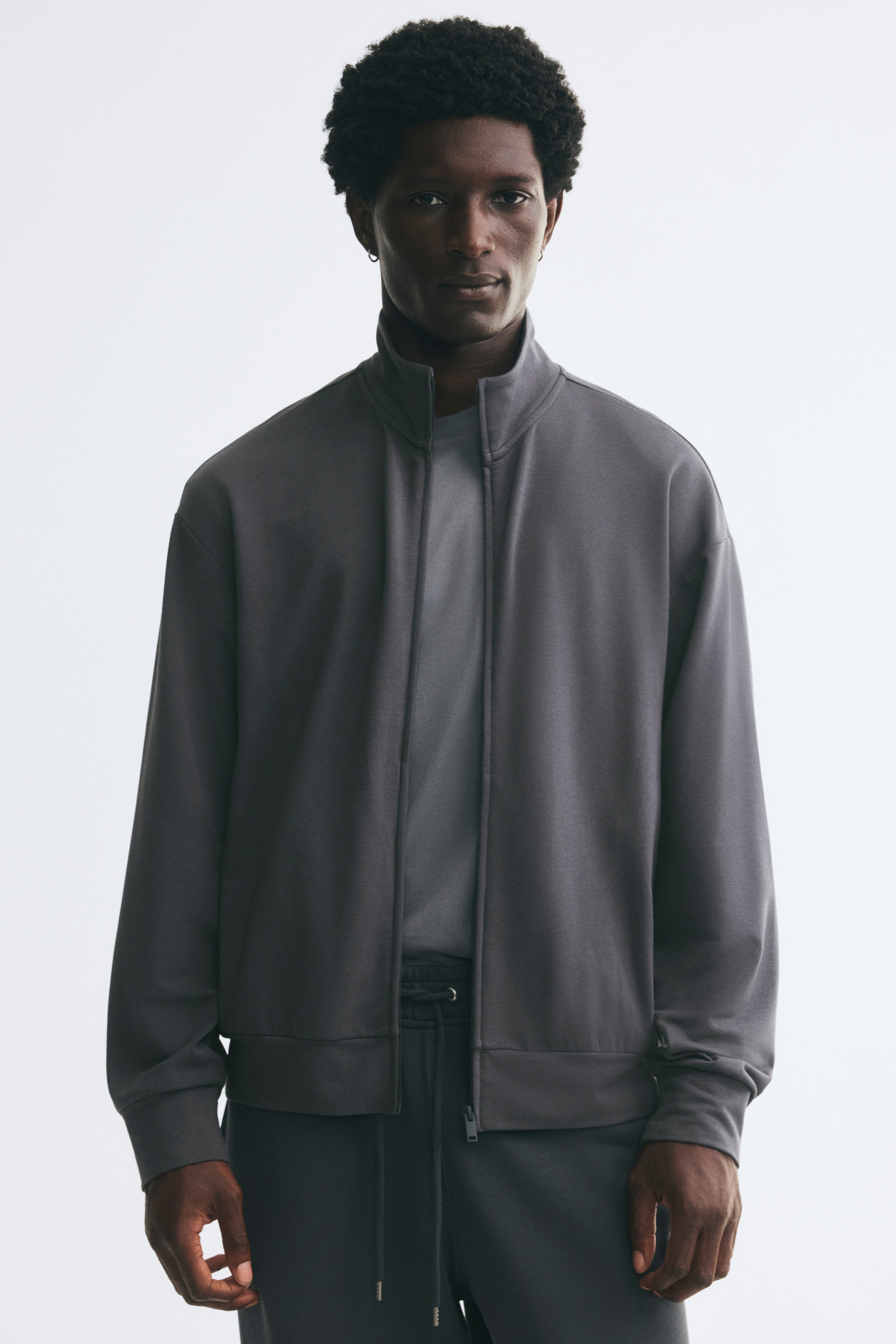 A dark grey sweat jacket is facing the front, showcasing a relaxed fit with a stand collar and a full-length front zipper. The jacket is featuring dropped shoulders, long sleeves with wide cuffs, and a wide hem.