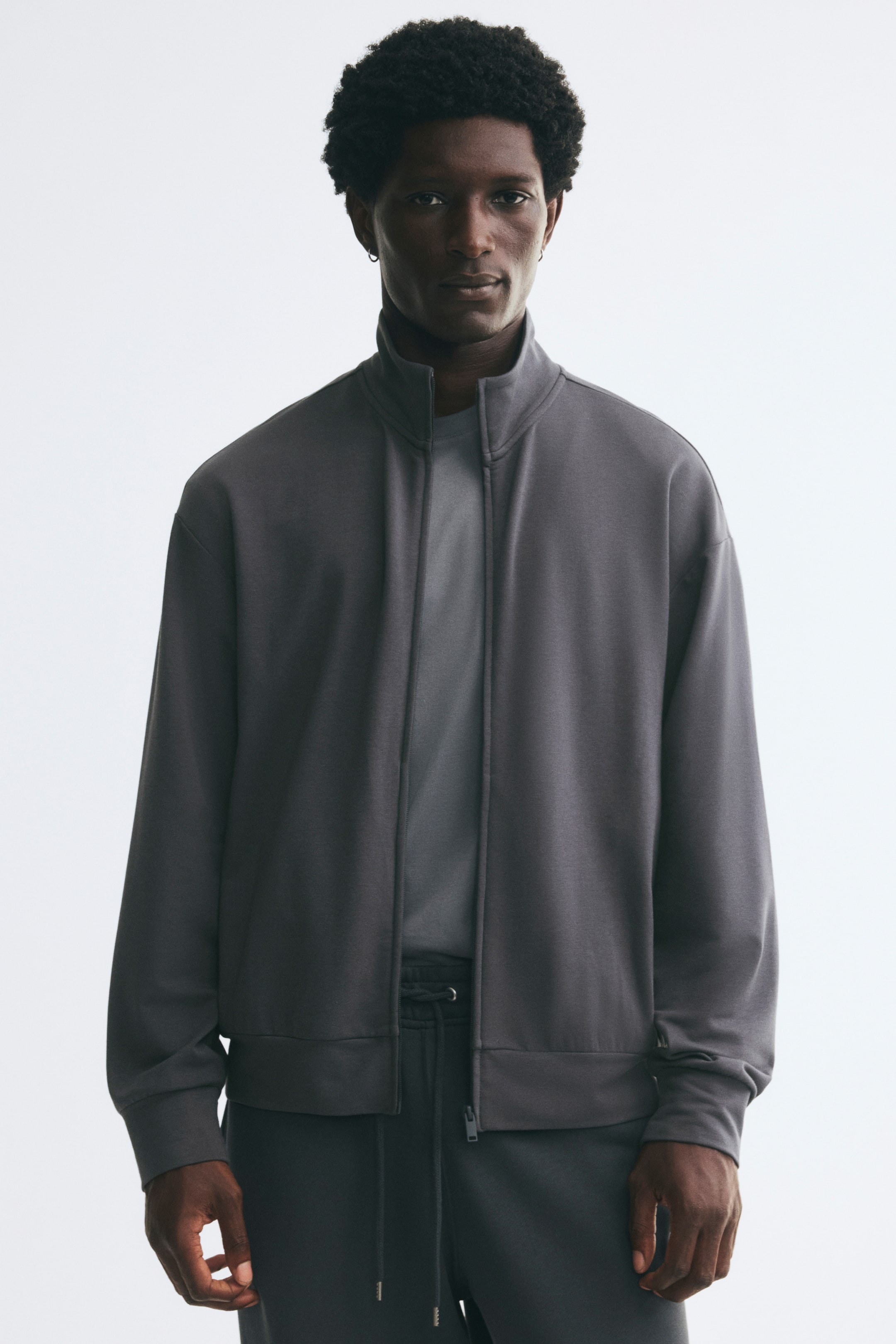 Voir une image plus grande: Une veste de survêtement gris foncé, dotée d’un col montant, d’épaules tombantes et d’une fermeture éclair à l’avant, est portée par le mannequin. La veste est stylée ouverte par-dessus un t-shirt gris plus clair et associée à un pantalon de survêtement gris foncé assorti avec cordon de serrage.