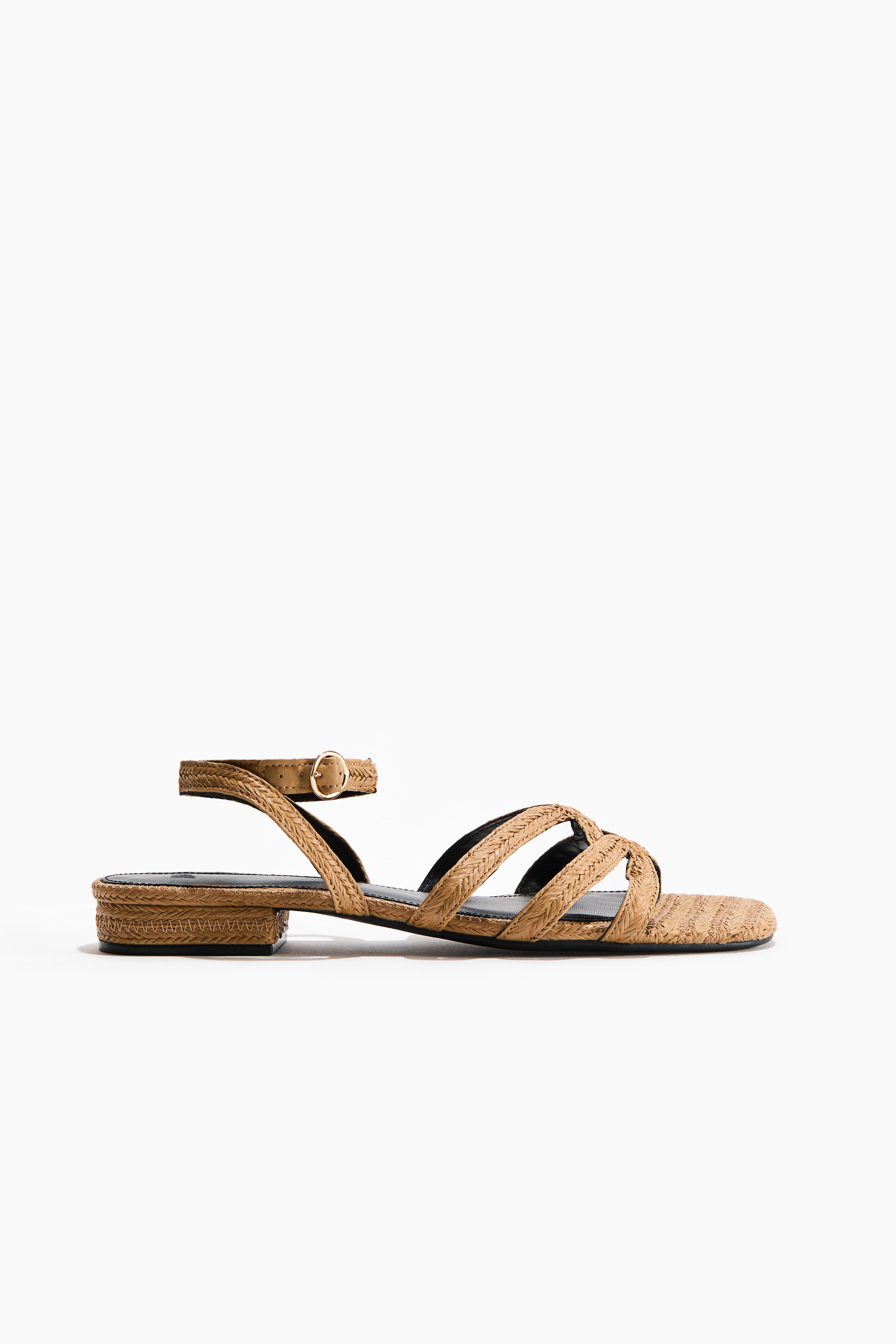 Sandaler i flettet strå med avrundede tær, smale fotremmer og en justerbar ankelrem med metallspenne bæres av modellen.