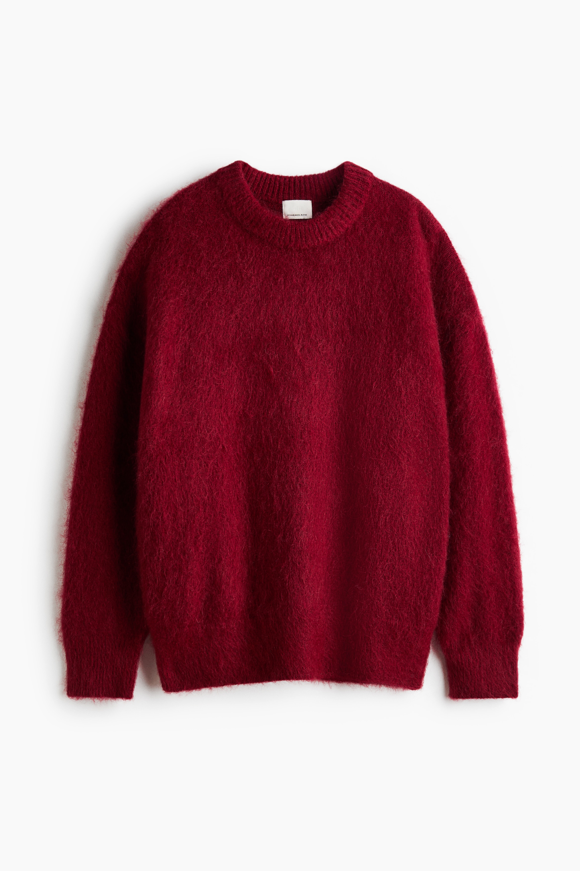 A red, fuzzy mohair-blend knit jumper with a ribbed round neckline is being worn by the model, with a matching red jumper draped over their shoulders. The jumper is layered over a white shirt, and the model is standing with hands placed in the pockets of dark trousers.