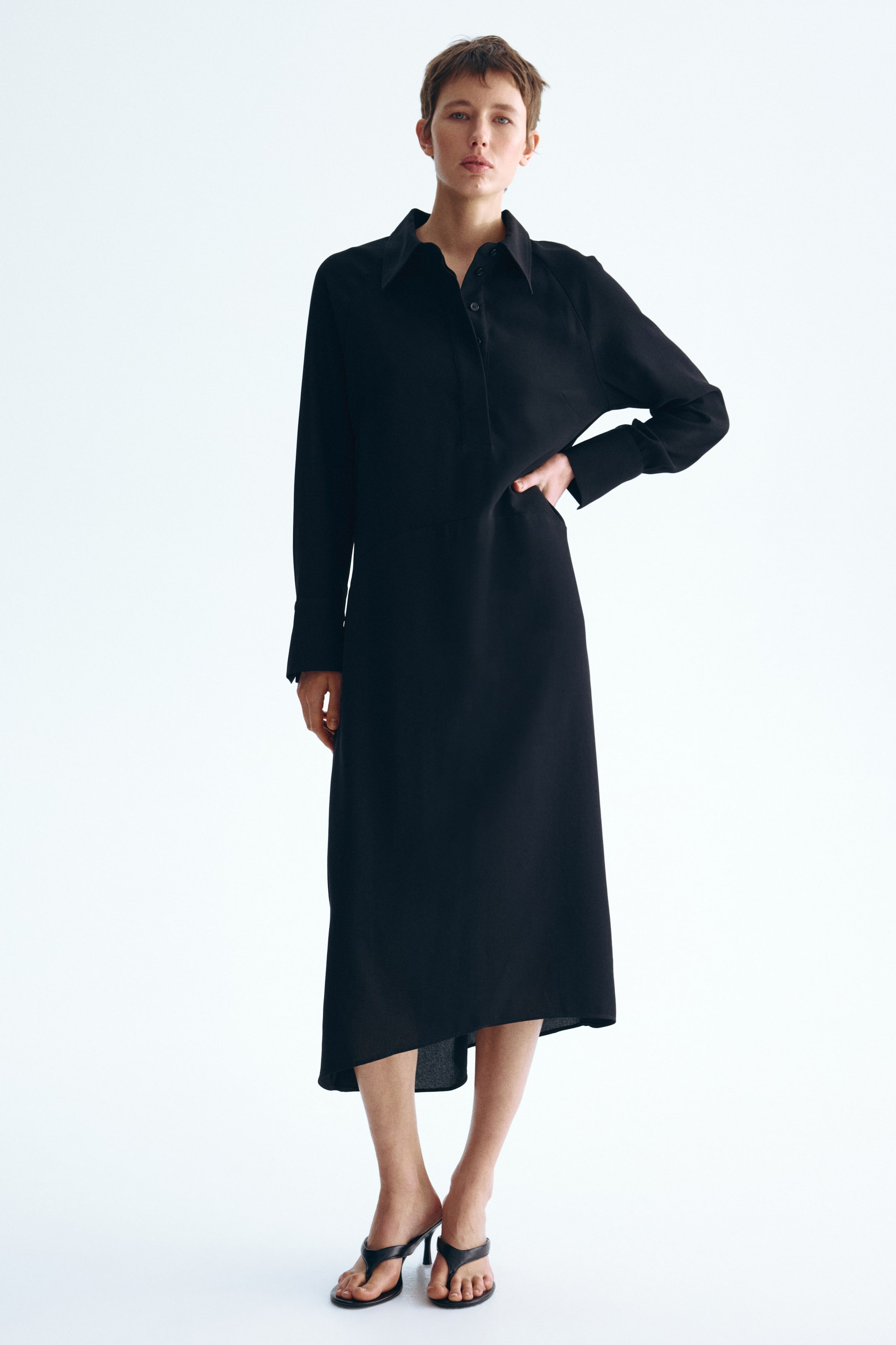 View larger image: A black midi-length shirt dress is displayed from a front angle, featuring a pointed collar, a half-button placket, and long cuffed sleeves. The dress is designed with a relaxed fit, an inseam pocket on the right side, and an asymmetrical hemline.