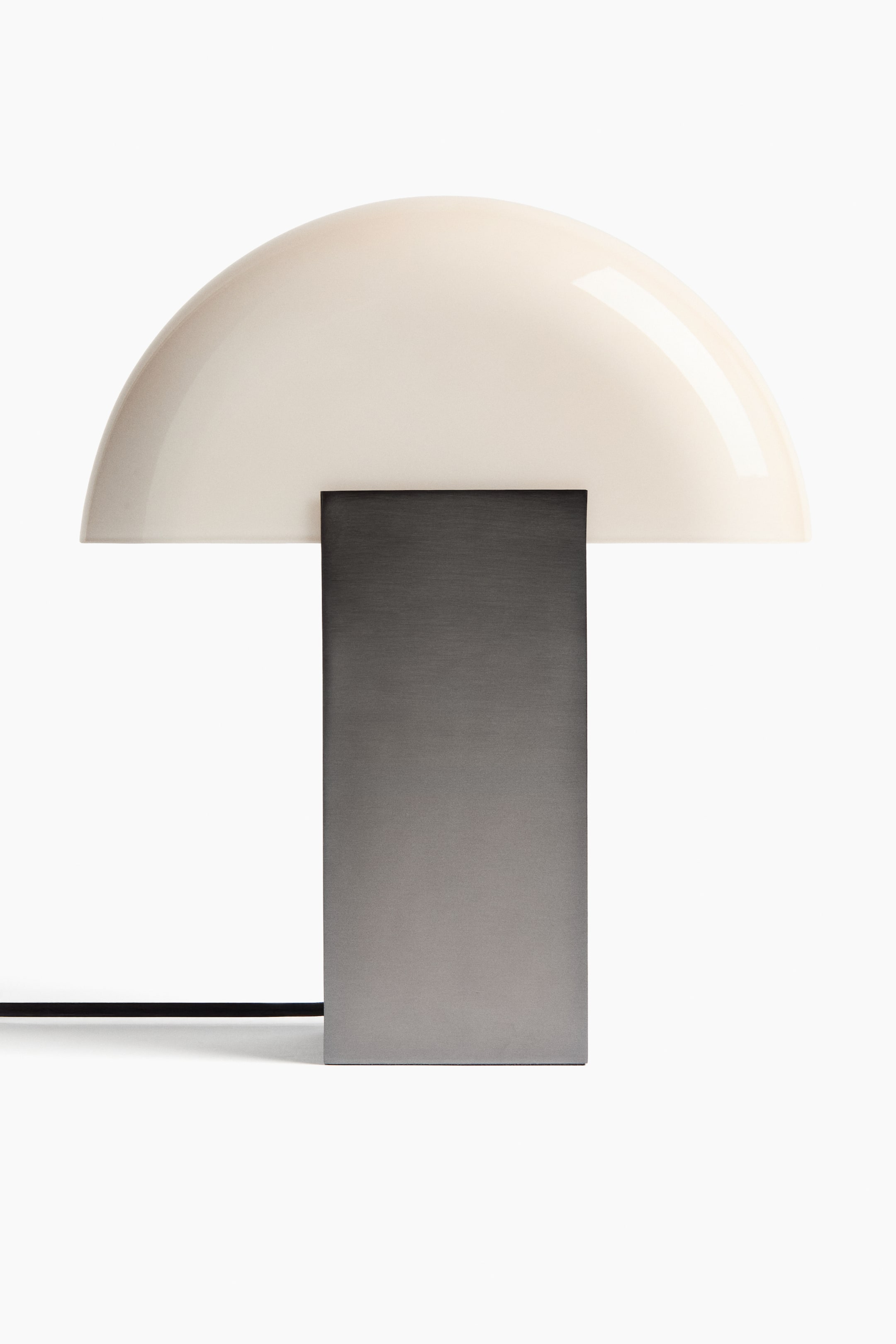 View larger image: A table lamp features a large, off-white, dome-shaped shade, supported by a rectangular dark grey metal base. A black power cord extends horizontally from the left side of the base.