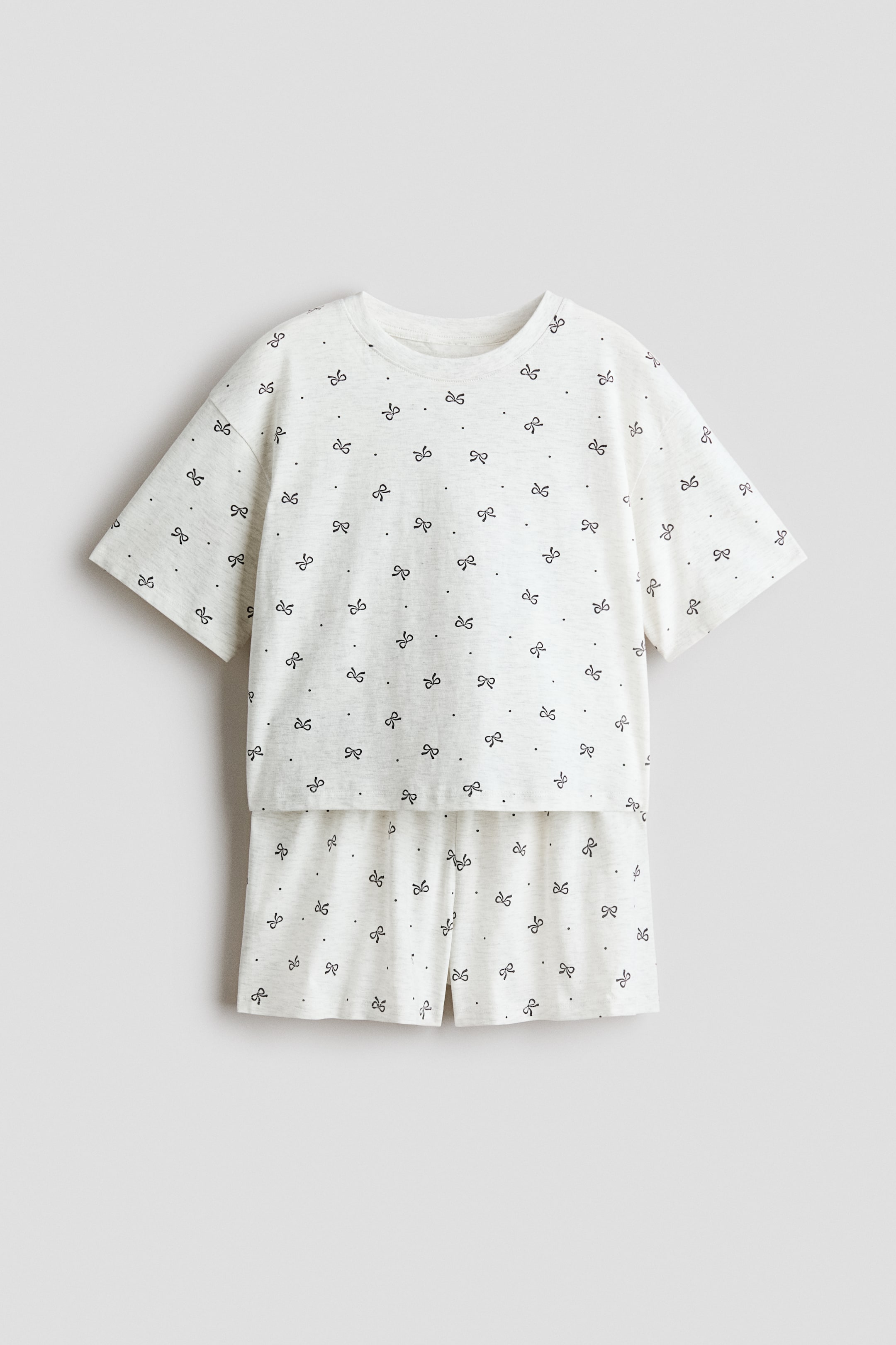 拡大画像を表示: 2点セットは、ライトカラーのTシャツとショートパンツで構成されており、どちらも小さな黒いリボンと小さな黒いドットの総柄が特徴です。半袖のTシャツはクルーネックデザインで、マッチングショートパンツはシンプルな裾でリラックスフィットを提供しています。