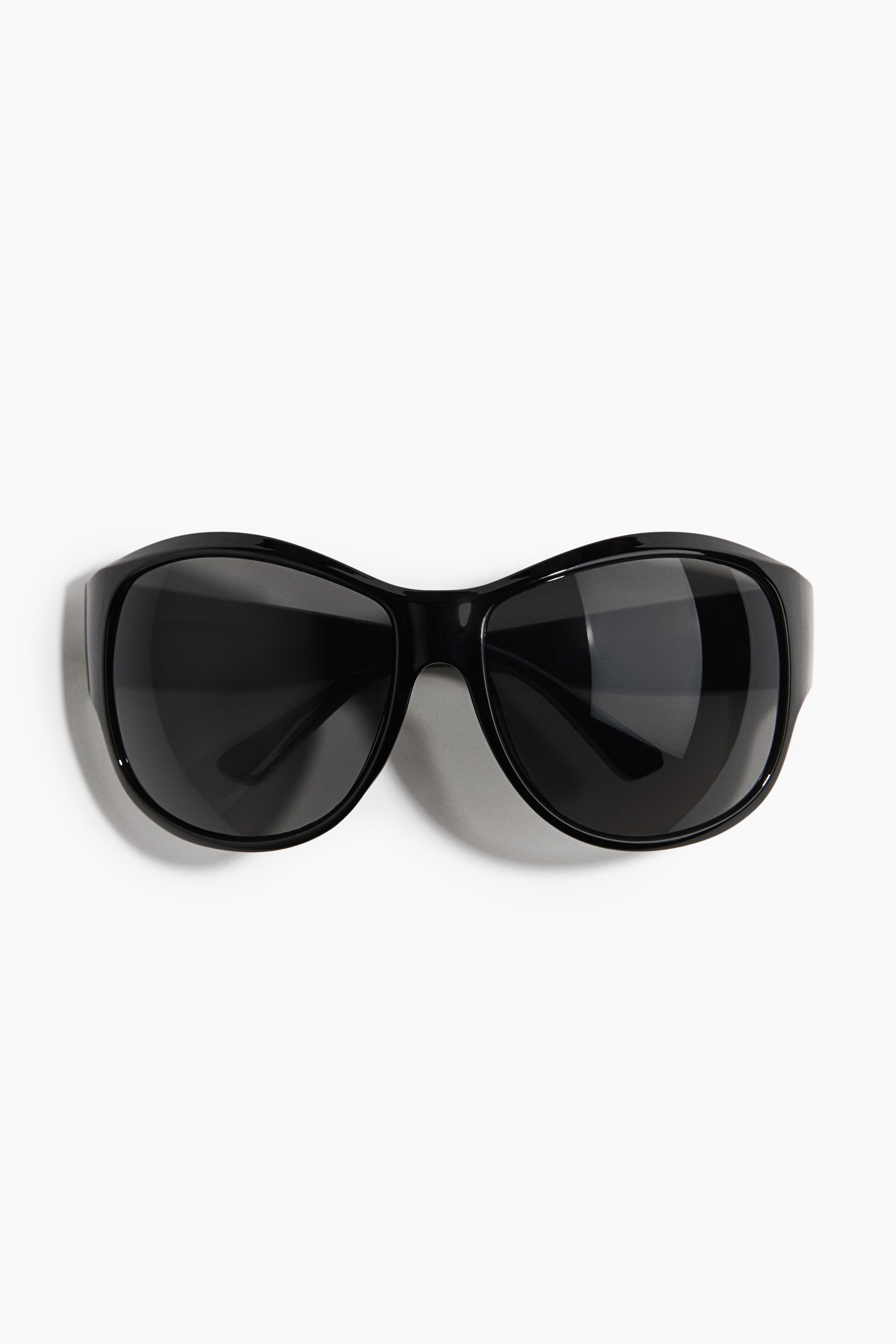 View larger image: Visor sunglasses featuring glossy black plastic frames and integrated sidepieces are lying flat, facing forwards. The sunglasses feature dark-tinted, curved plastic lenses in a wrap-around shape.