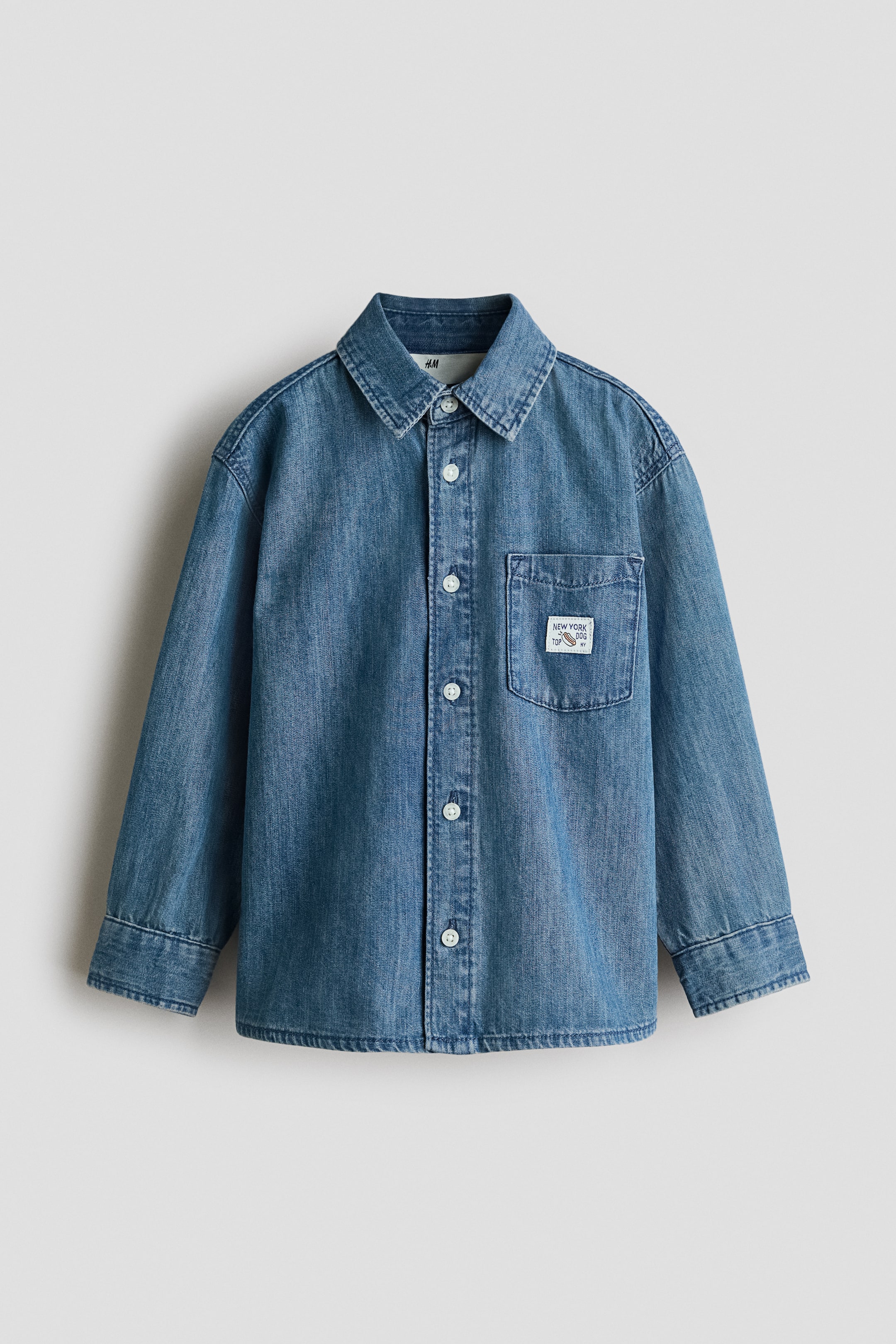 View larger image: A light blue denim shirt is hanging front facing, featuring a pointed collar, a full button-down front with white buttons, and long sleeves ending in buttoned cuffs. A patch pocket is appearing on the left chest, displaying a white label with "NEW YORK DOG" text and a hot dog graphic.