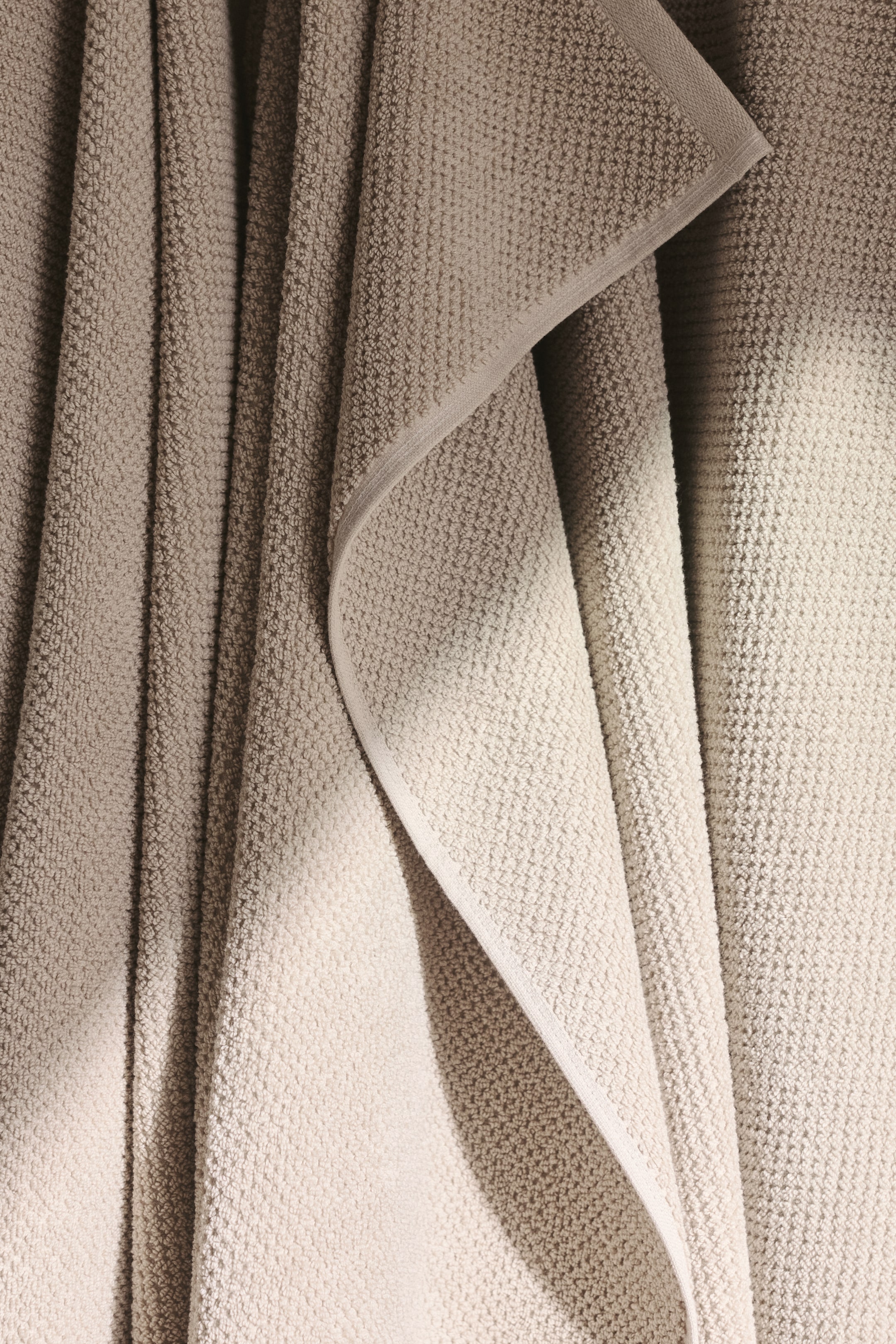 Visa större bild: En ljus beige frottéhandduk hänger, avslöjande sin texturerade yta och en distinkt vikt kant med ett slätt band.